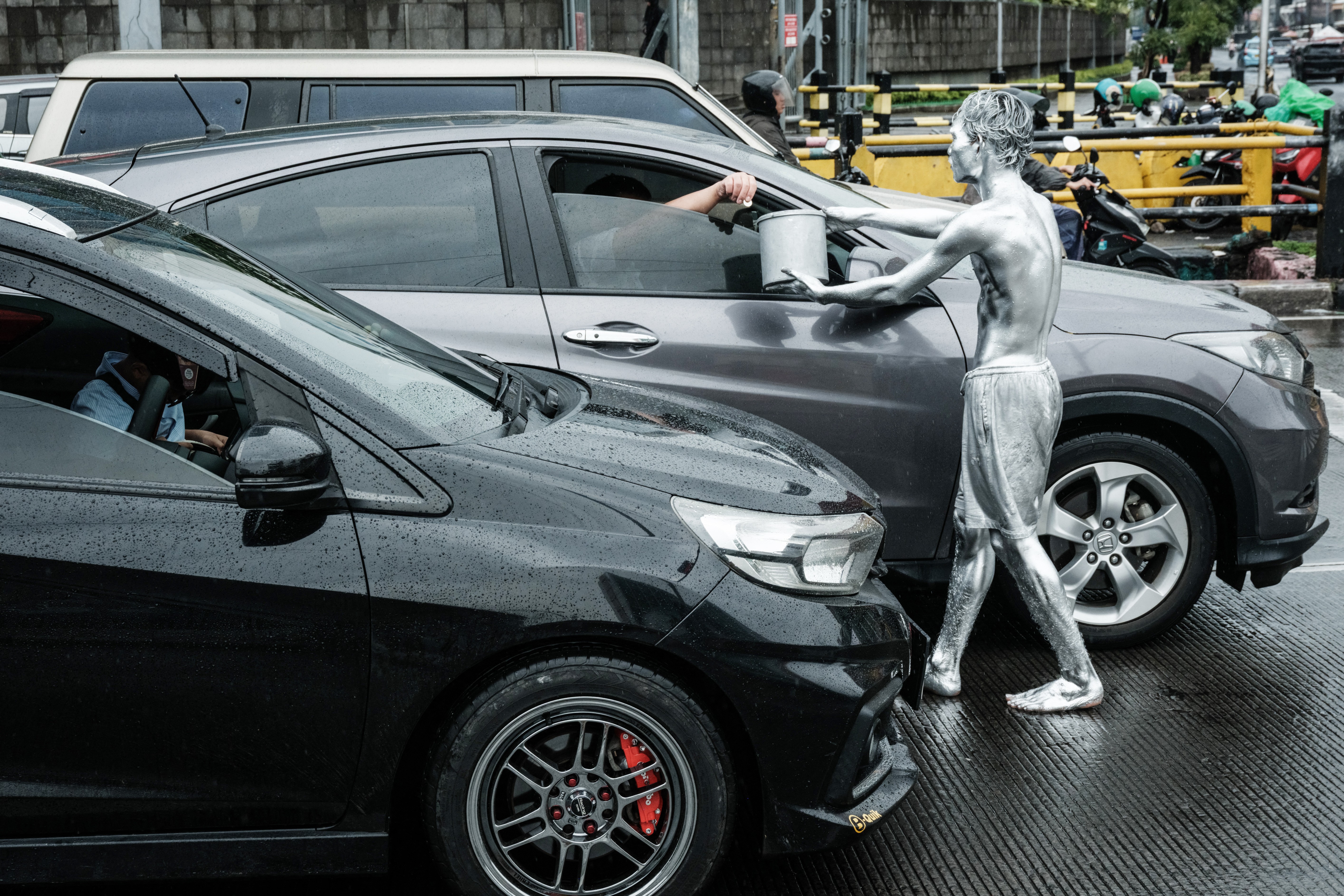 A street performer covered in silver paint holds out a bucket for tips beside a car.