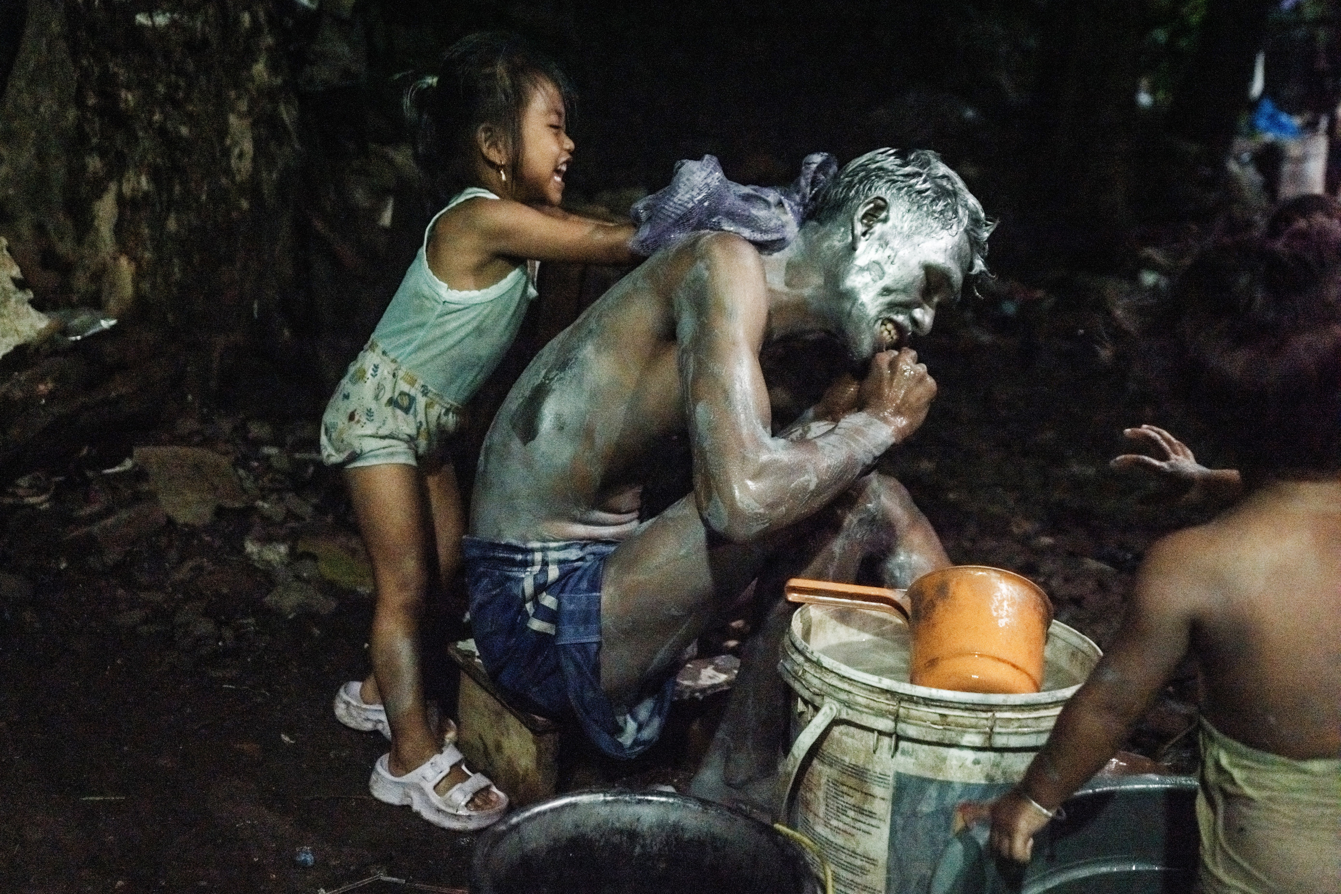 A man kneels down while a young girl playfully scrubs his back with soap and water.