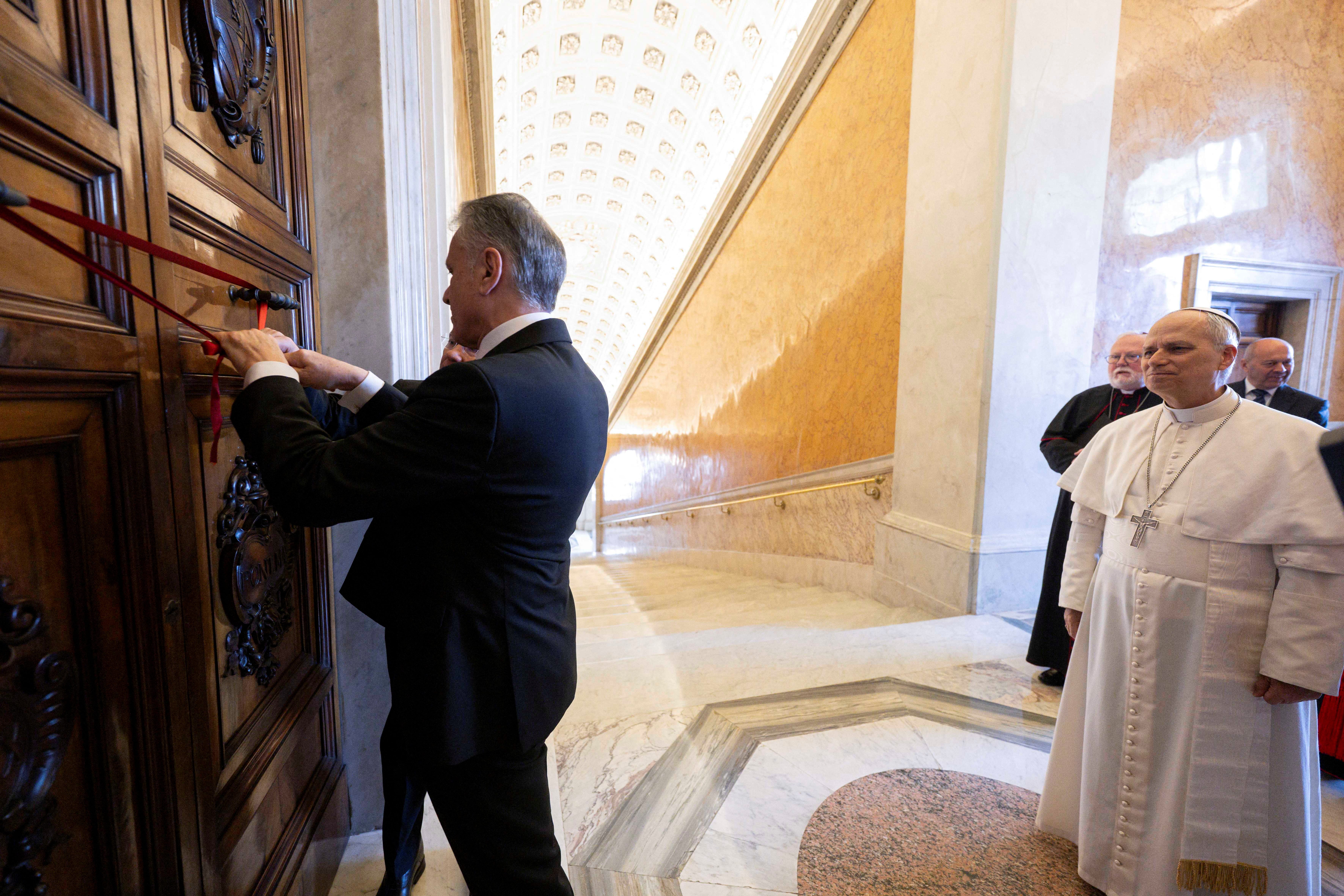 A man in a suit removes a red ribbon and seal from a door as Pope Leo XIV looks on