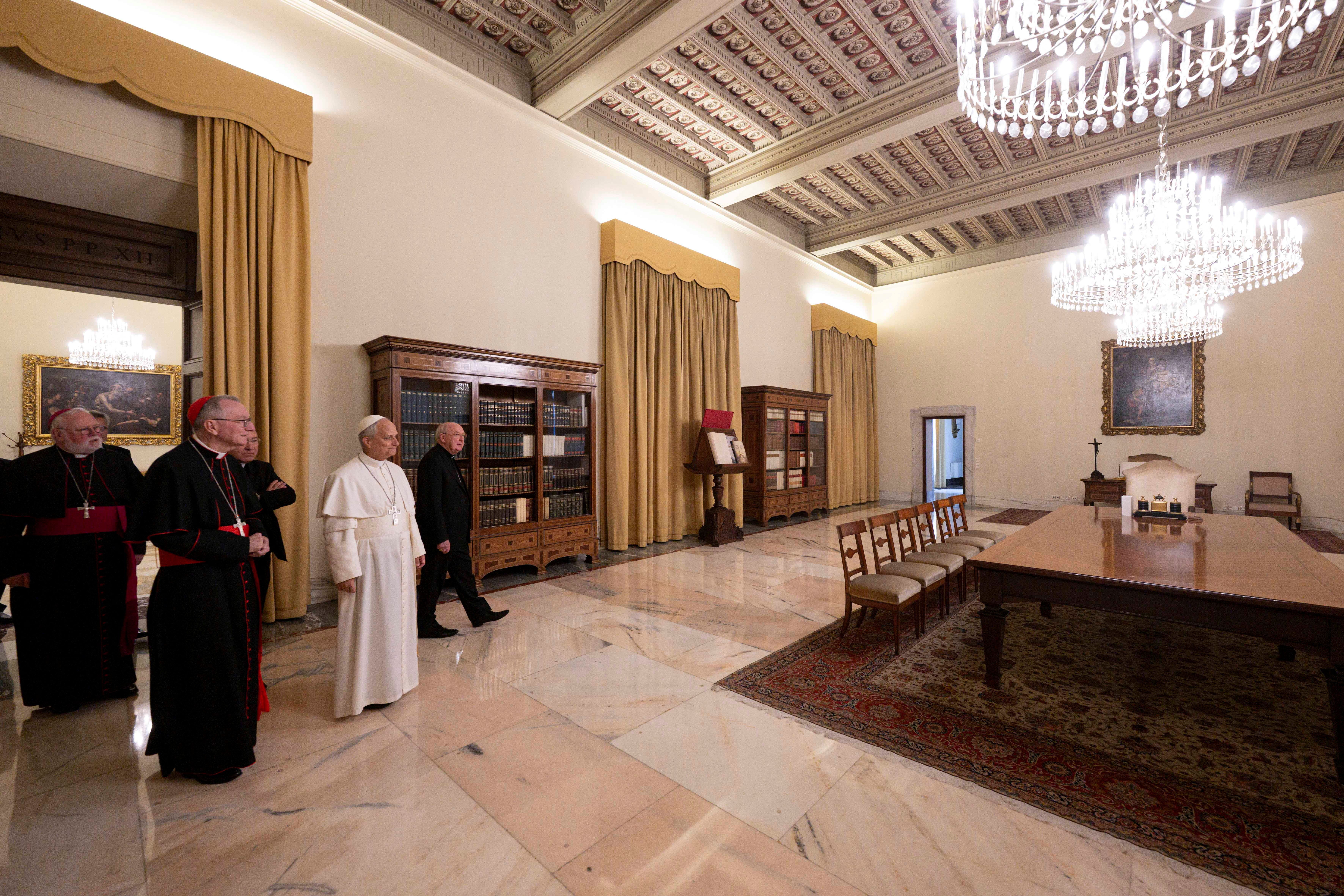 Pope Loe and others stand in a room with large chandeliers and a table and chairs
