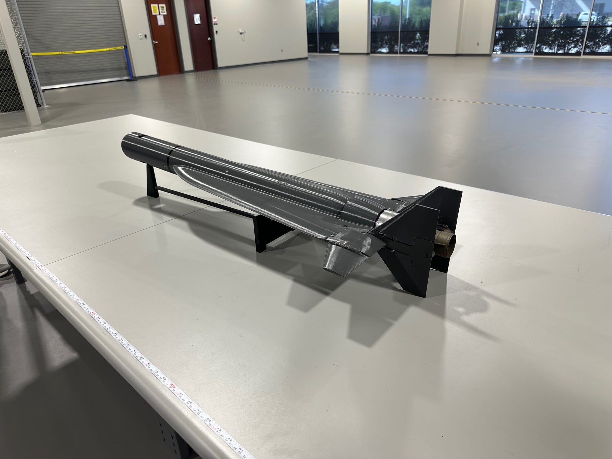 A black loitering munition is seen from the left-back side sitting on a white table in an office space.