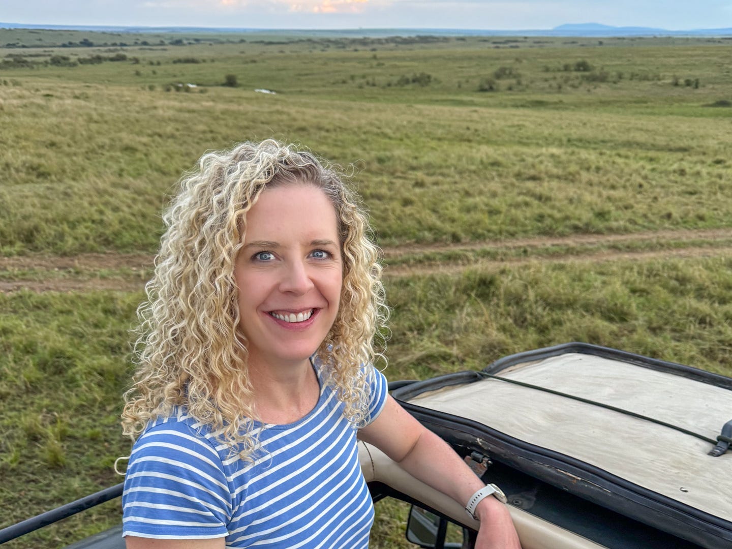 Blonde, curly haried woman standing in a car in a blue and white striped shirt.