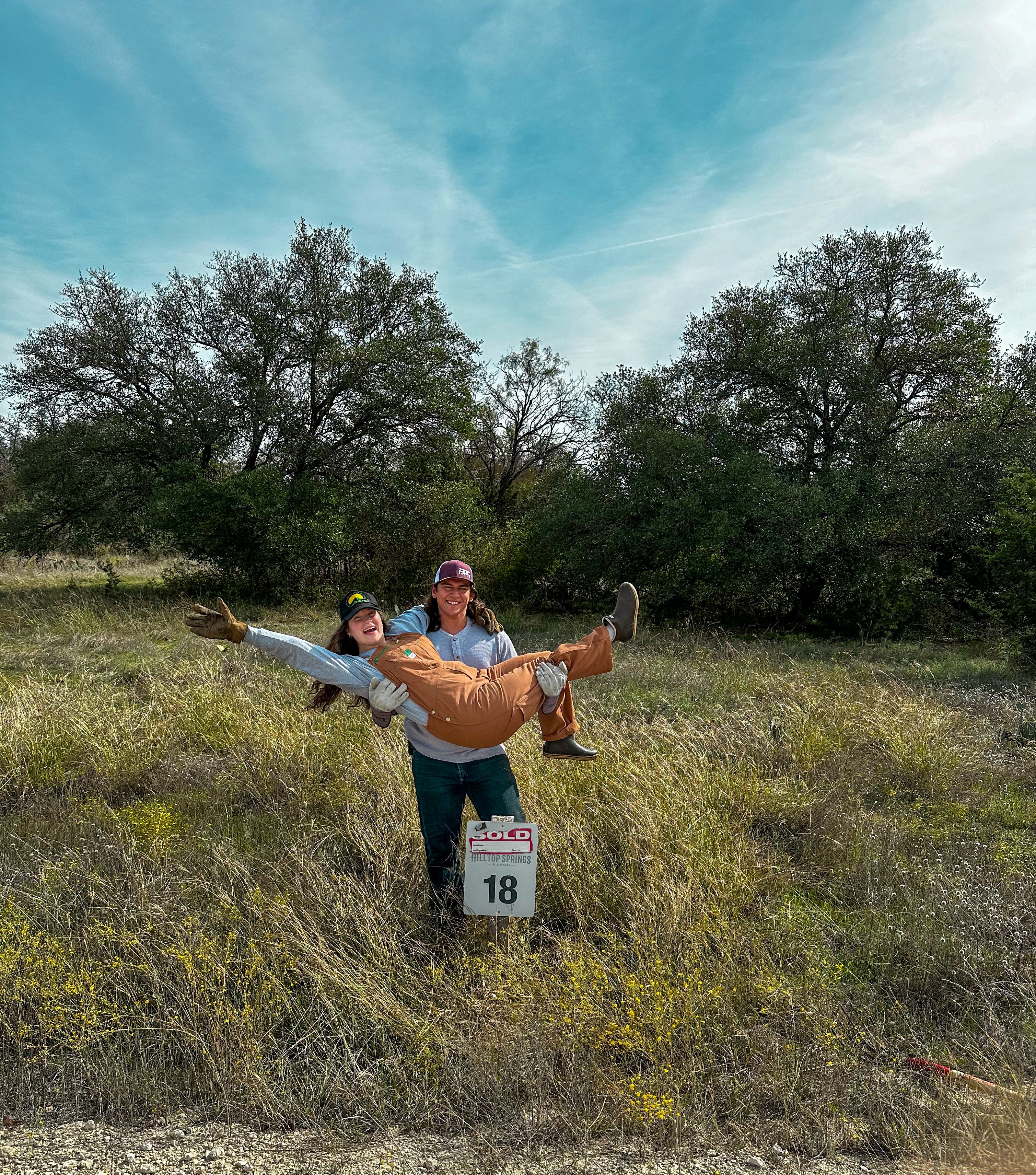 A man carries a woman in a field in front of a "sold" sign.