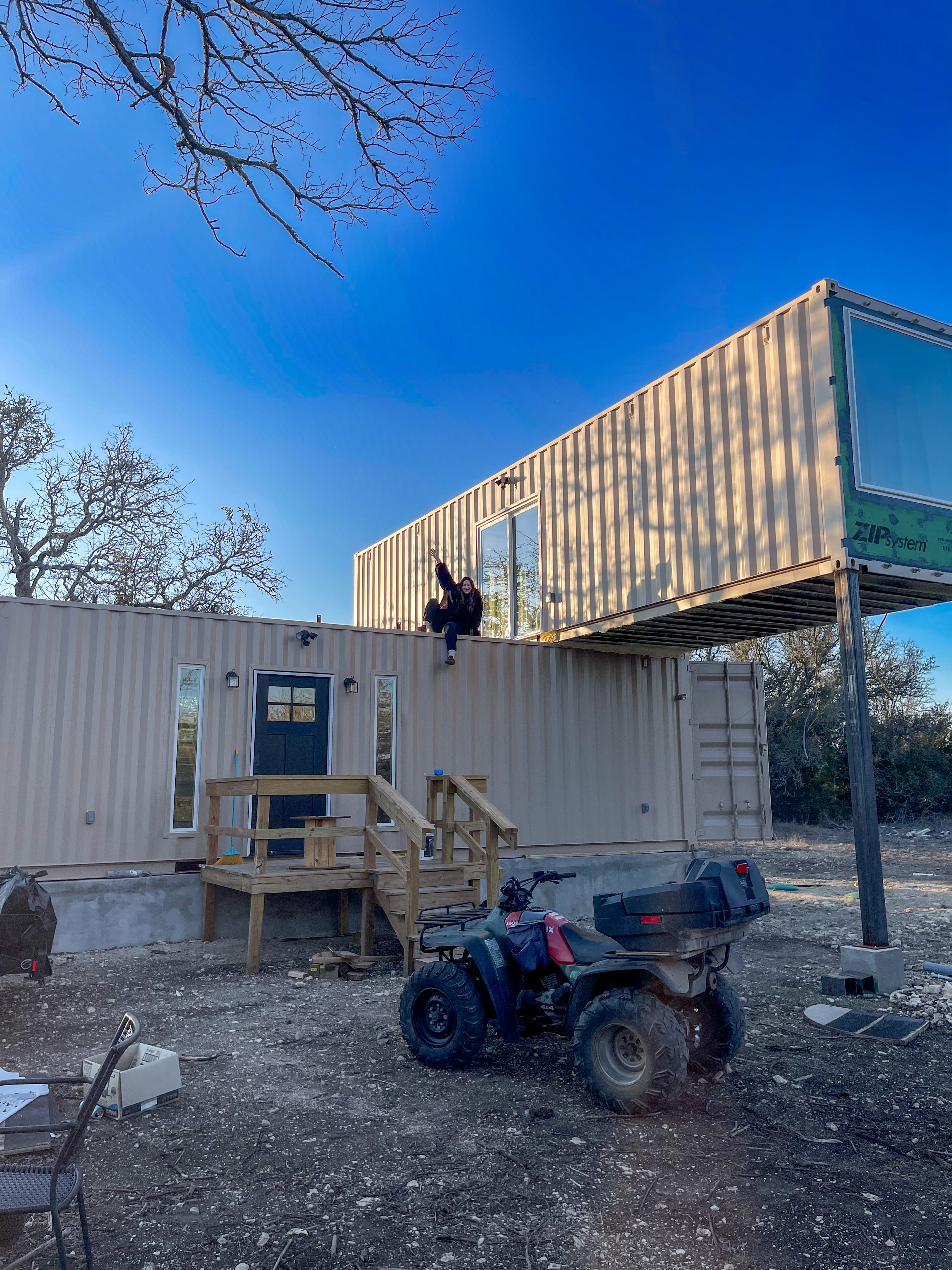 A photo of a shipping container home with two containers stacked together. A woman waves from the top of the shorter one.