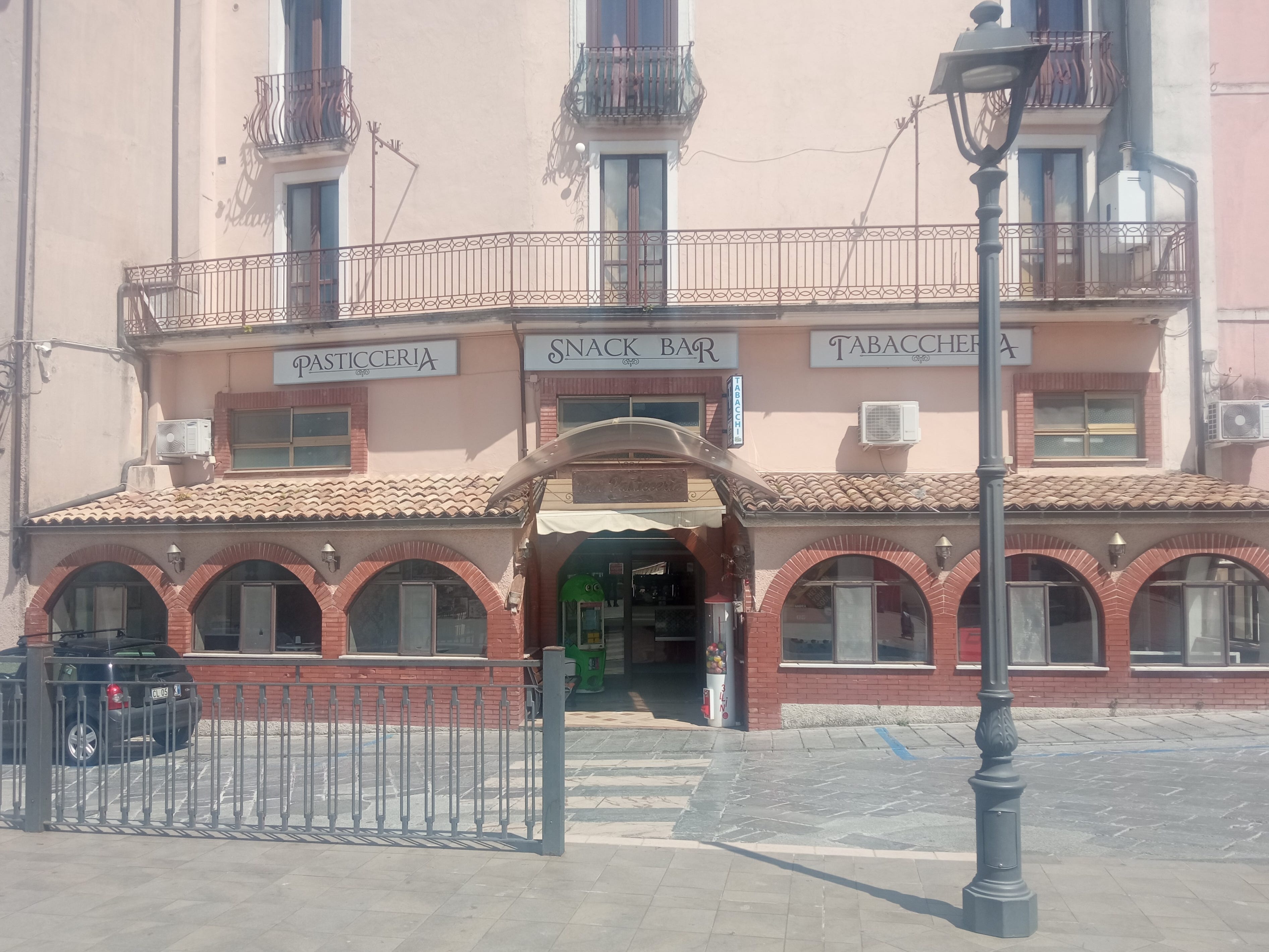 A wide shot of a building in Mormanno, Italy, with signs for "Pasticceria," "Snack Bar," and "Tabacchera."