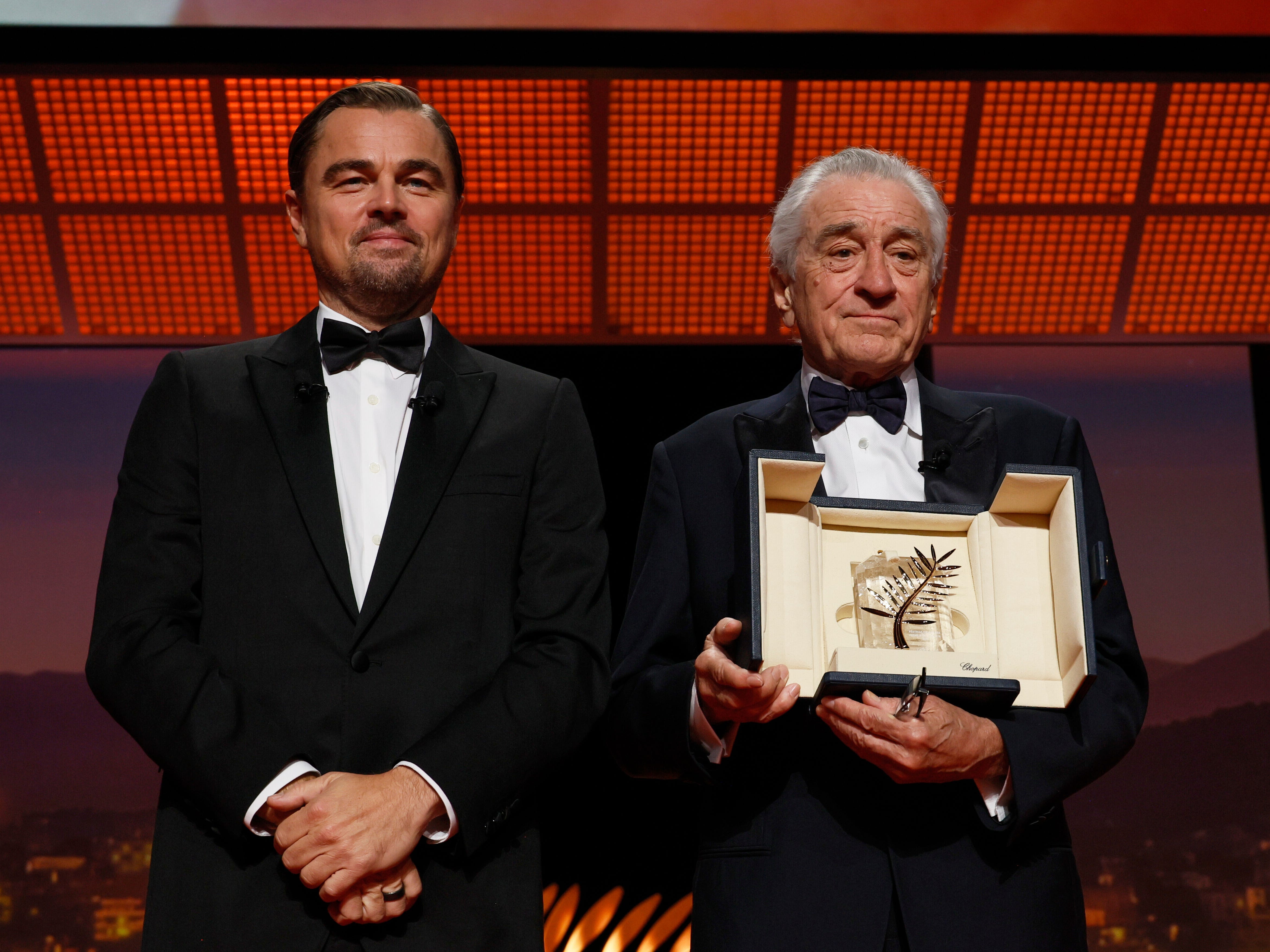 Two men in tuxedos. On the right, the middle-aged man has brown hair and a brown goatee. He's holding his hands in front of him, and there is a black ring on his middle finger on his left hand. On the right, an older man with gray hair holds a black box with a beige interior. There is a silver trophy shaped like a leaf inside.