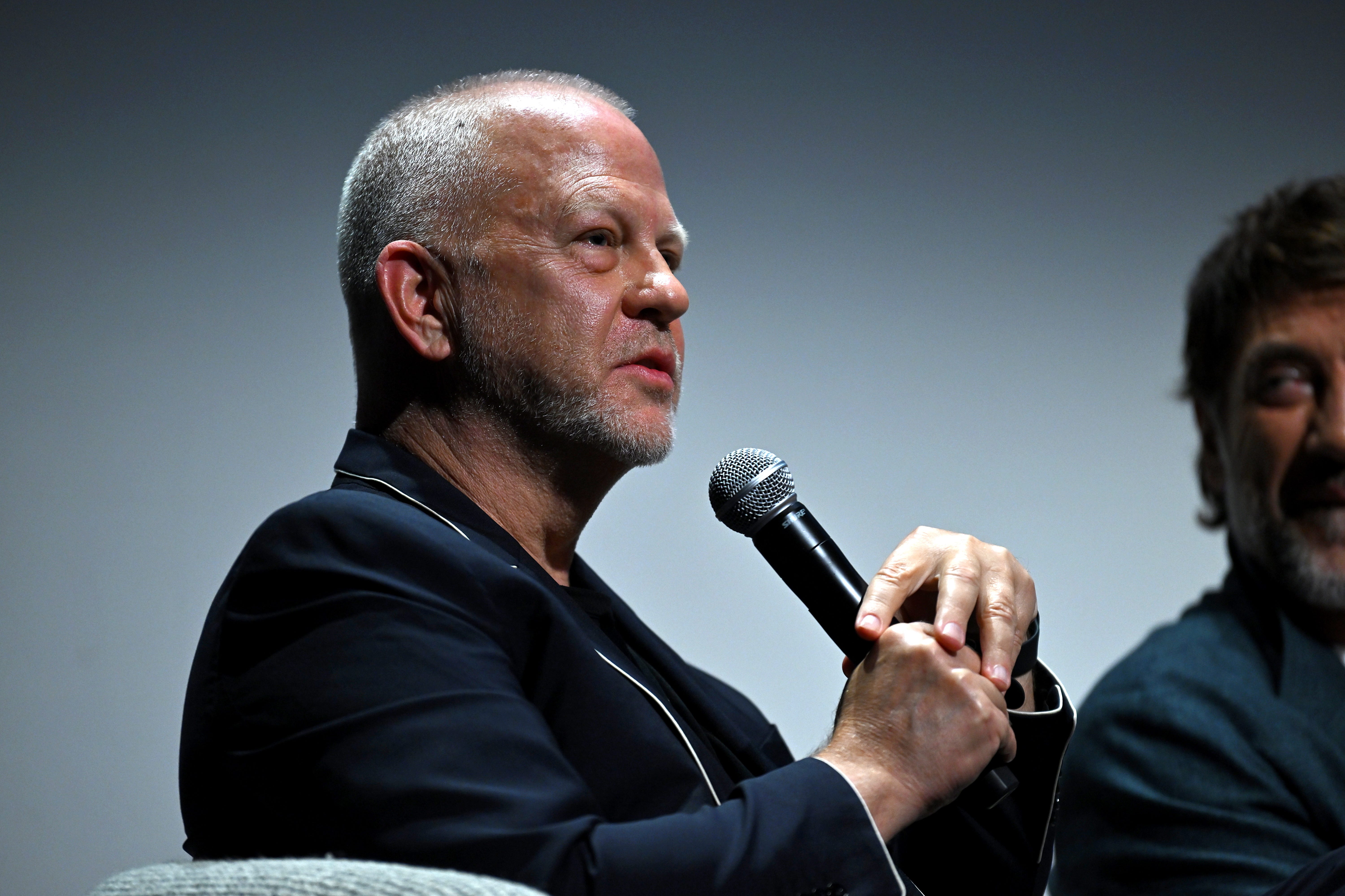 A man with short gray hair and stubble wearing a navy blue blazer with white piping. He's holding a microphone.