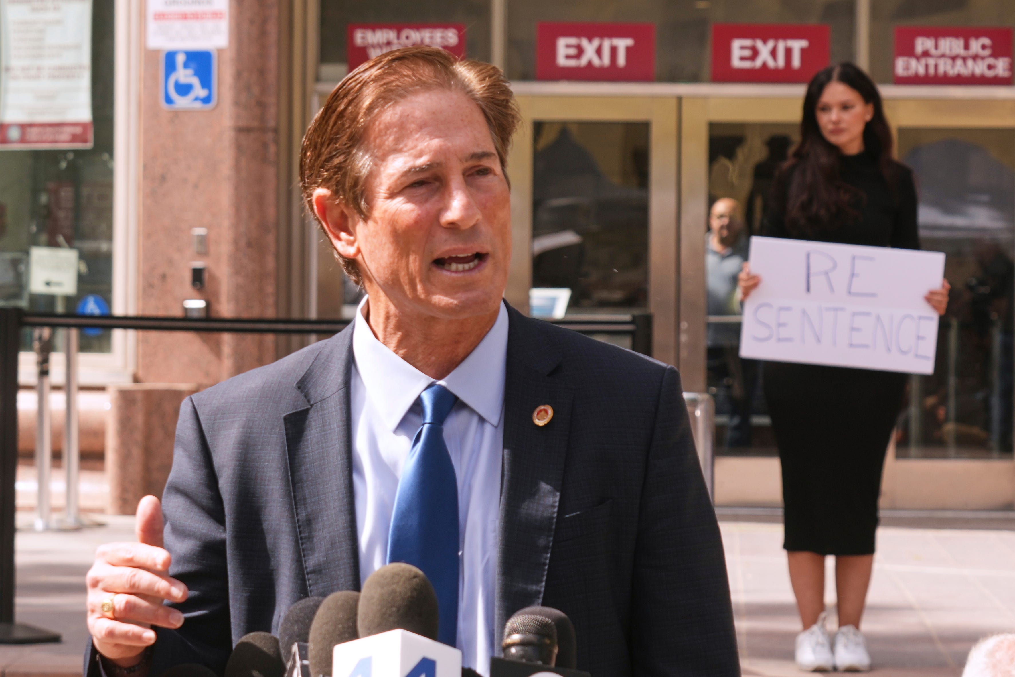 A photo of Nathan Hochman in a formally blue suit and tie, speaking into multiple microphones while standing in front of a building.