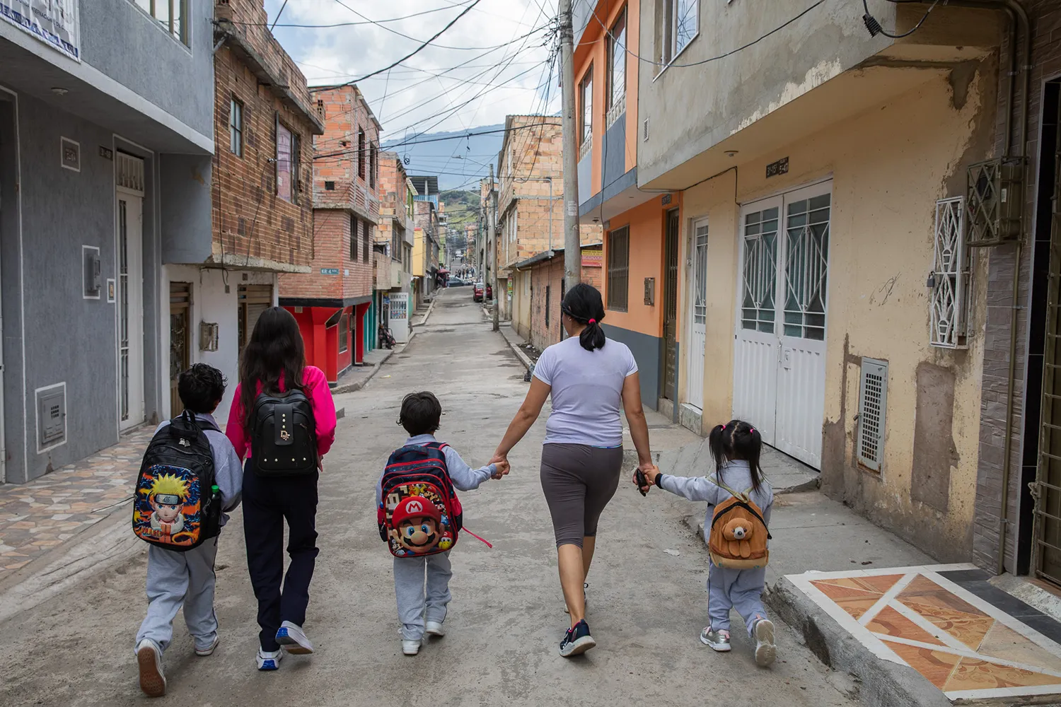 A mom and four children, waring backpacks, hold hands as they walk away down a street with buildings on either side.
