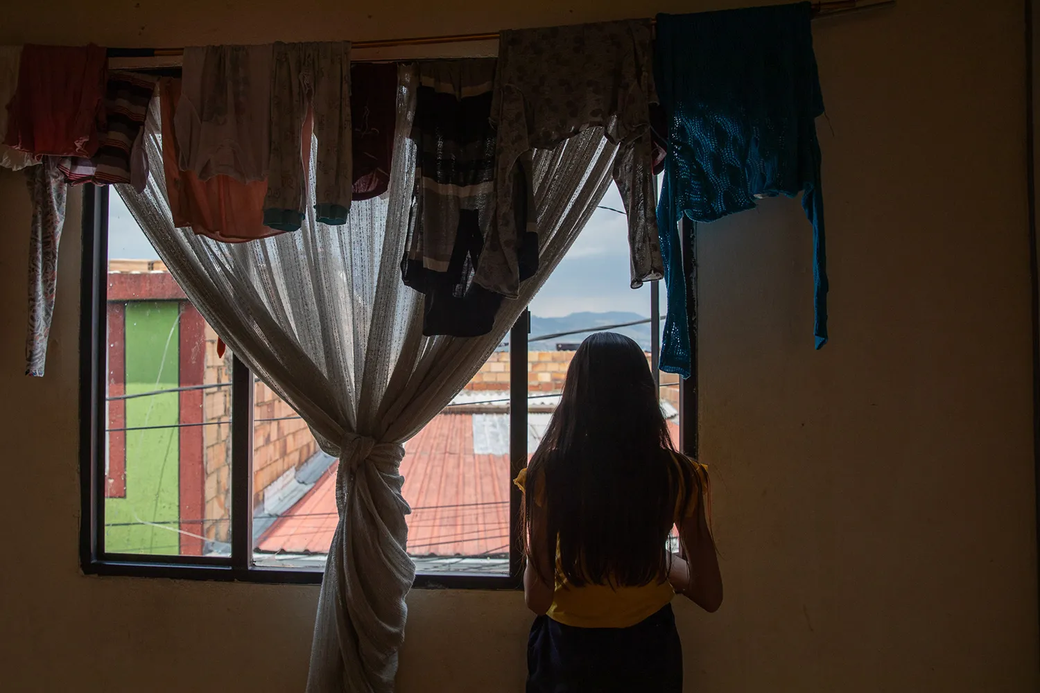A girl's silhouette is seen in a window. Rooftops and mountains are seen outside. A knotted curtain and clothes hang over a curtain rod.