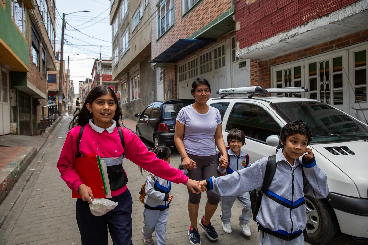 A woman holds hands with two young children in track suits in front of her are two older children holding hands. They walk past cars down a street with mid-rise buildings and power lines on either side.