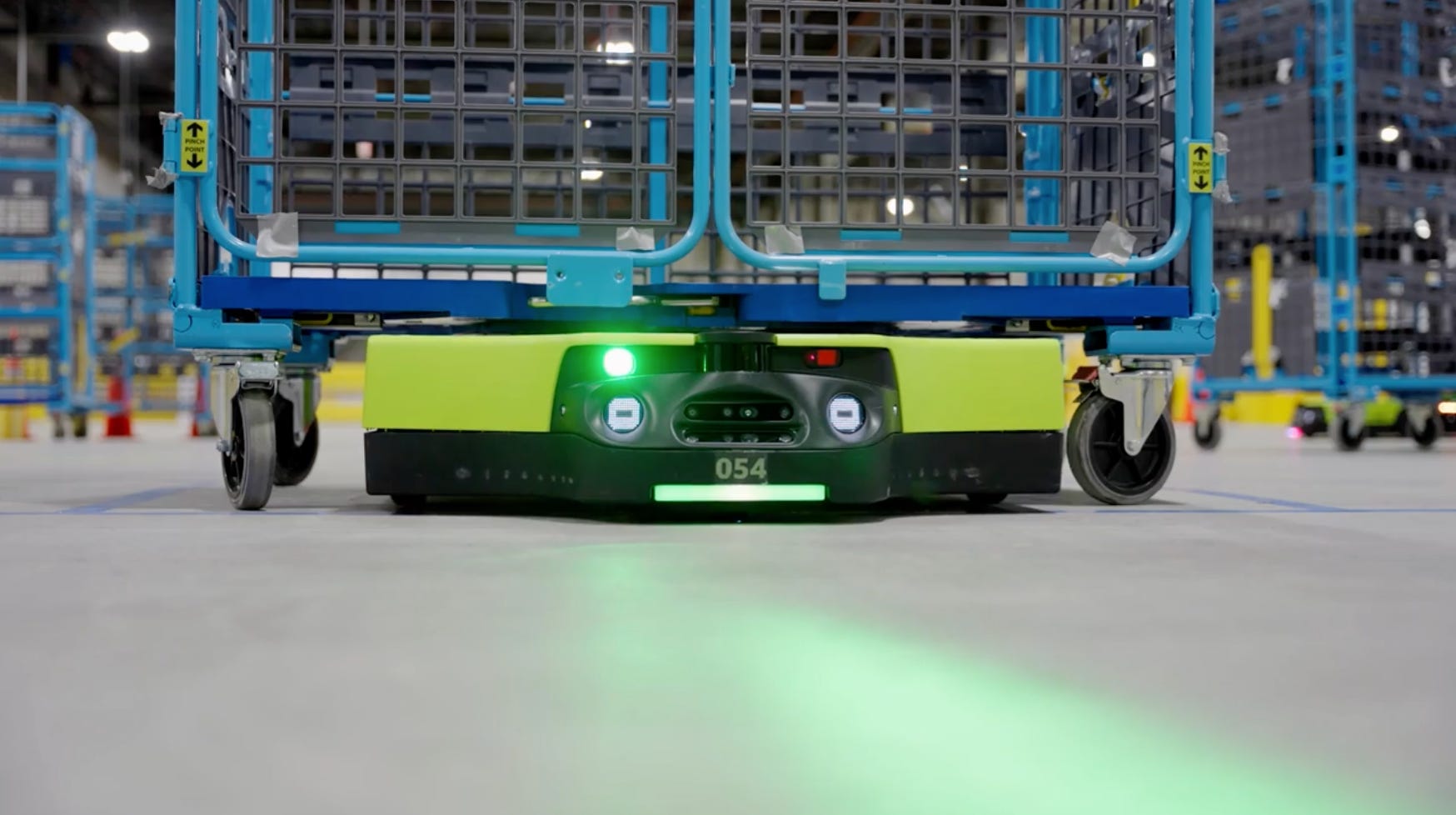 A green wheeled robot carries a large wheeled cage on its back.