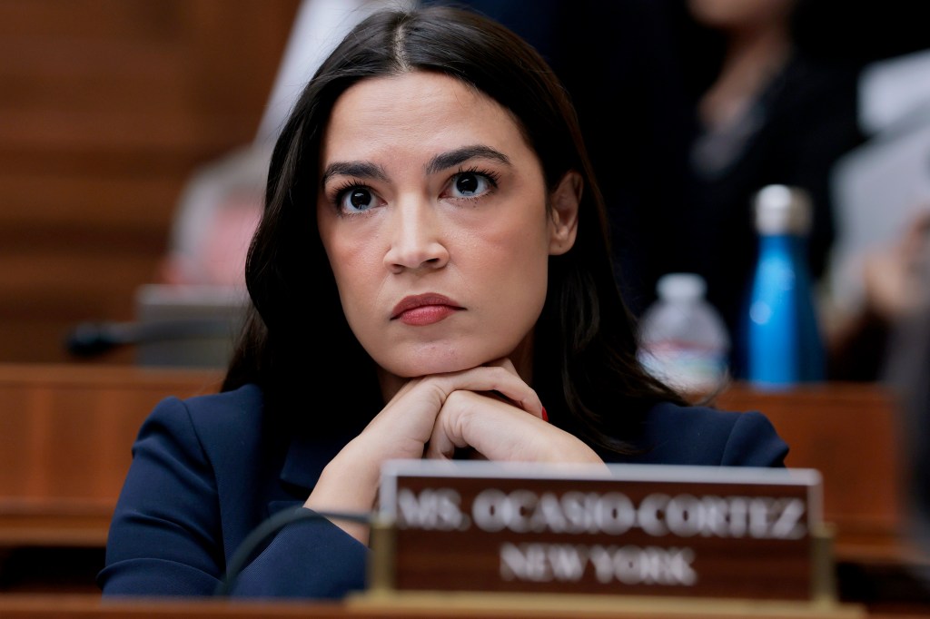 Rep. Alexandria Ocasio-Cortez attentively listening during a House Committee on Energy and Commerce meeting on Capitol Hill, with protesters visible in background