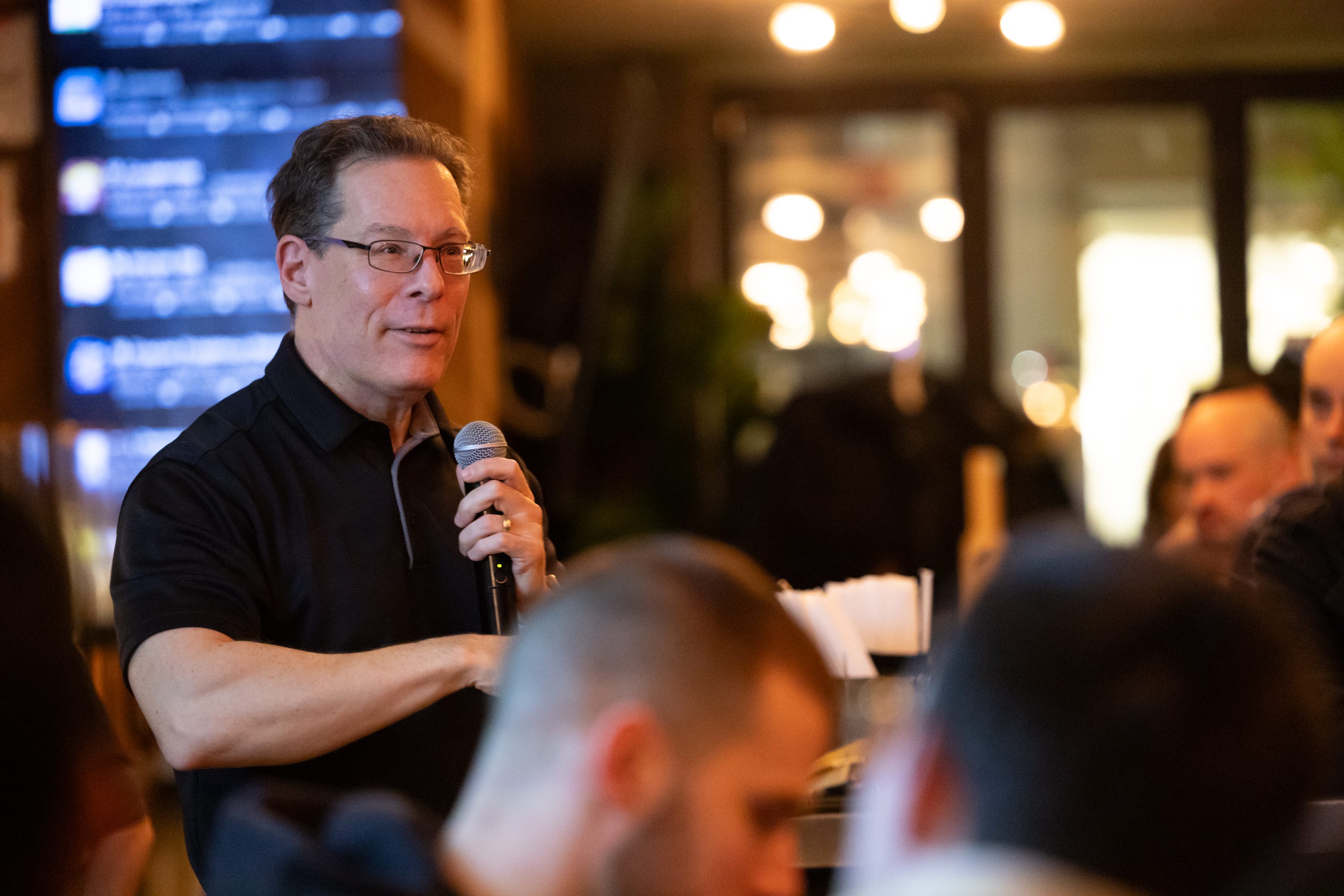 A man wearing glasses is holding a microphone and speaking to people inside an office space.