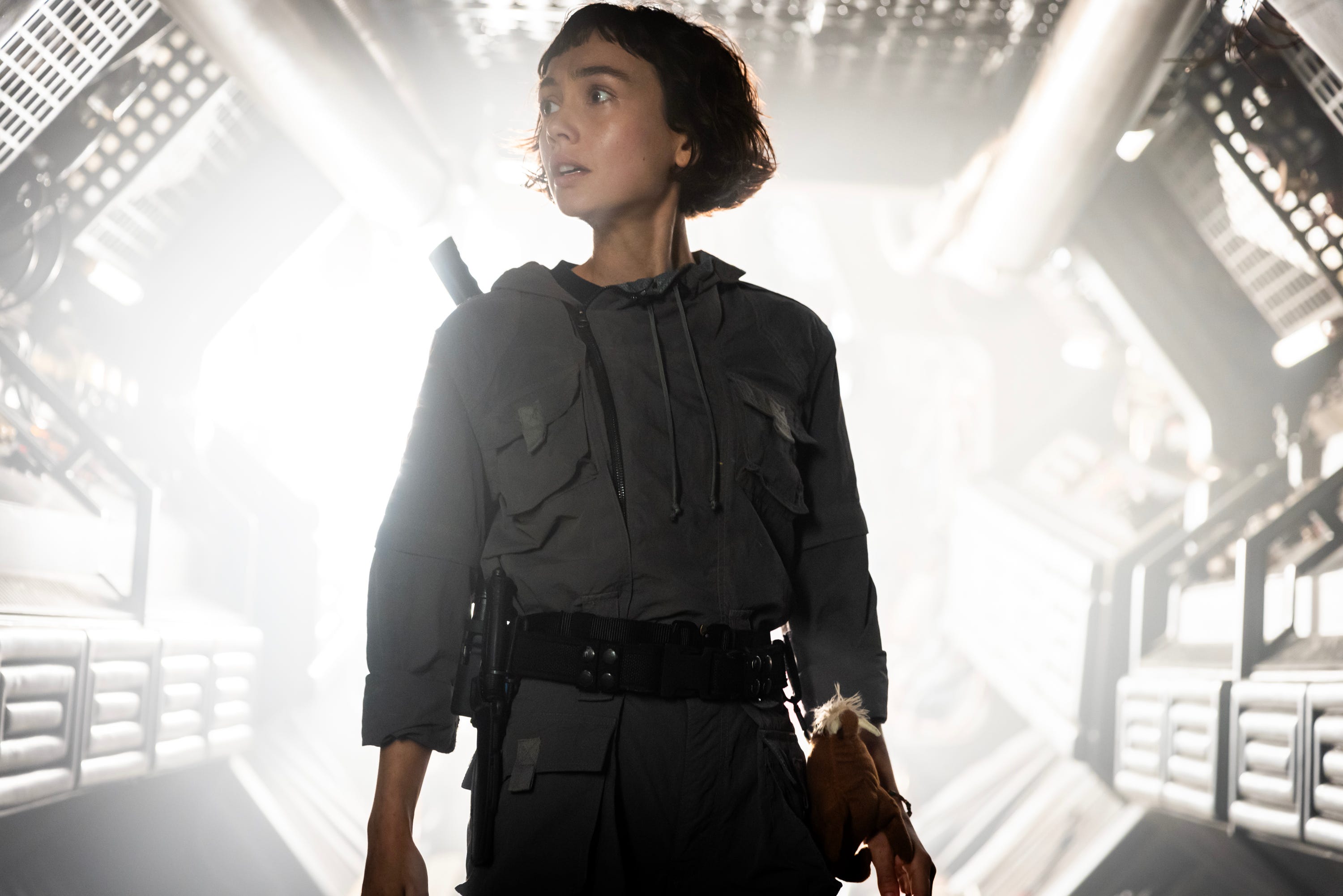 A woman with short brown hair looks to the left. She's wearing a gray boiler suit and has a black belt around her waist. There is also a cuddly toy strapped to her side.
