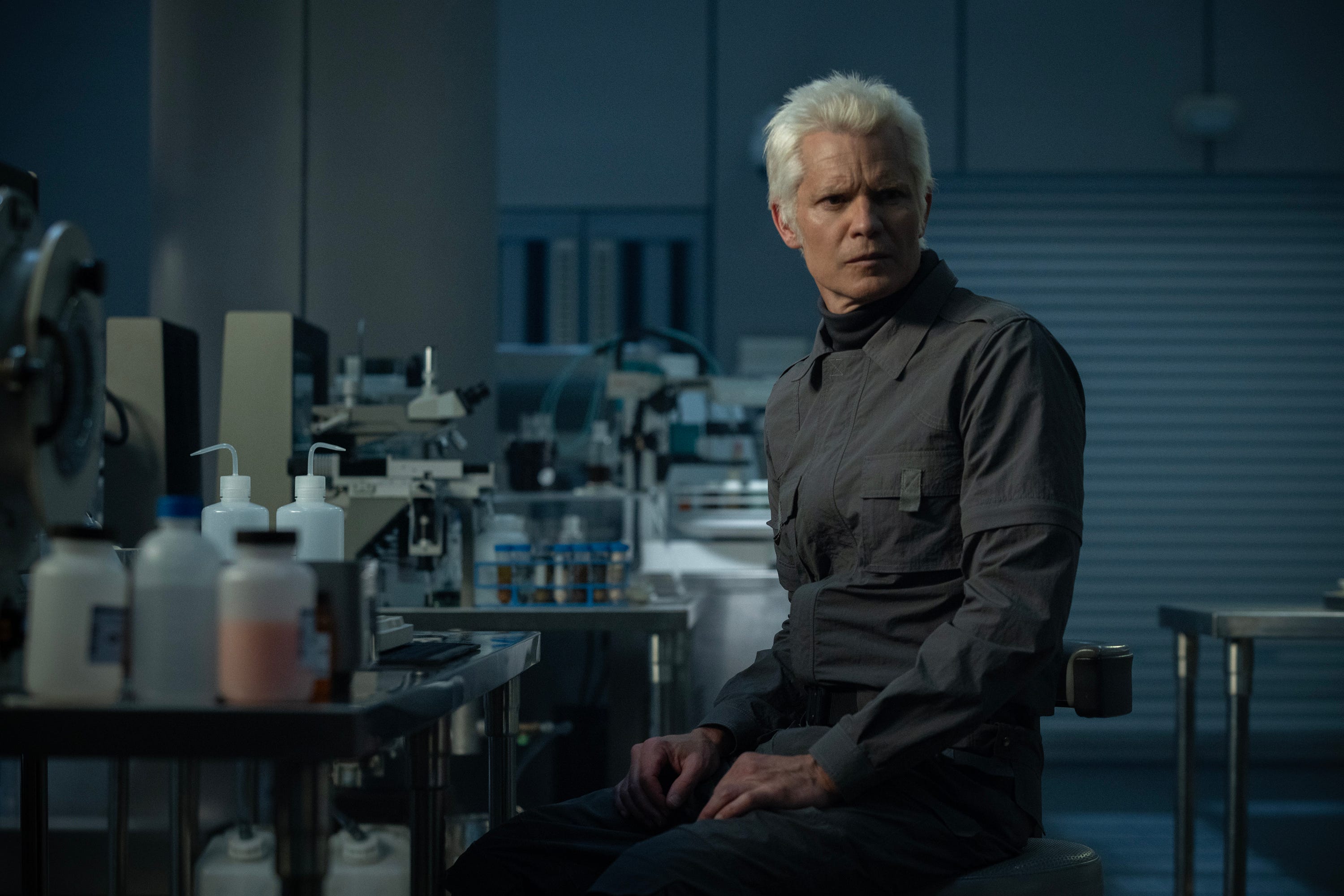 A man in. gray boilersuit has white hair. He's sitting at a table surrounded by scientific equipment.