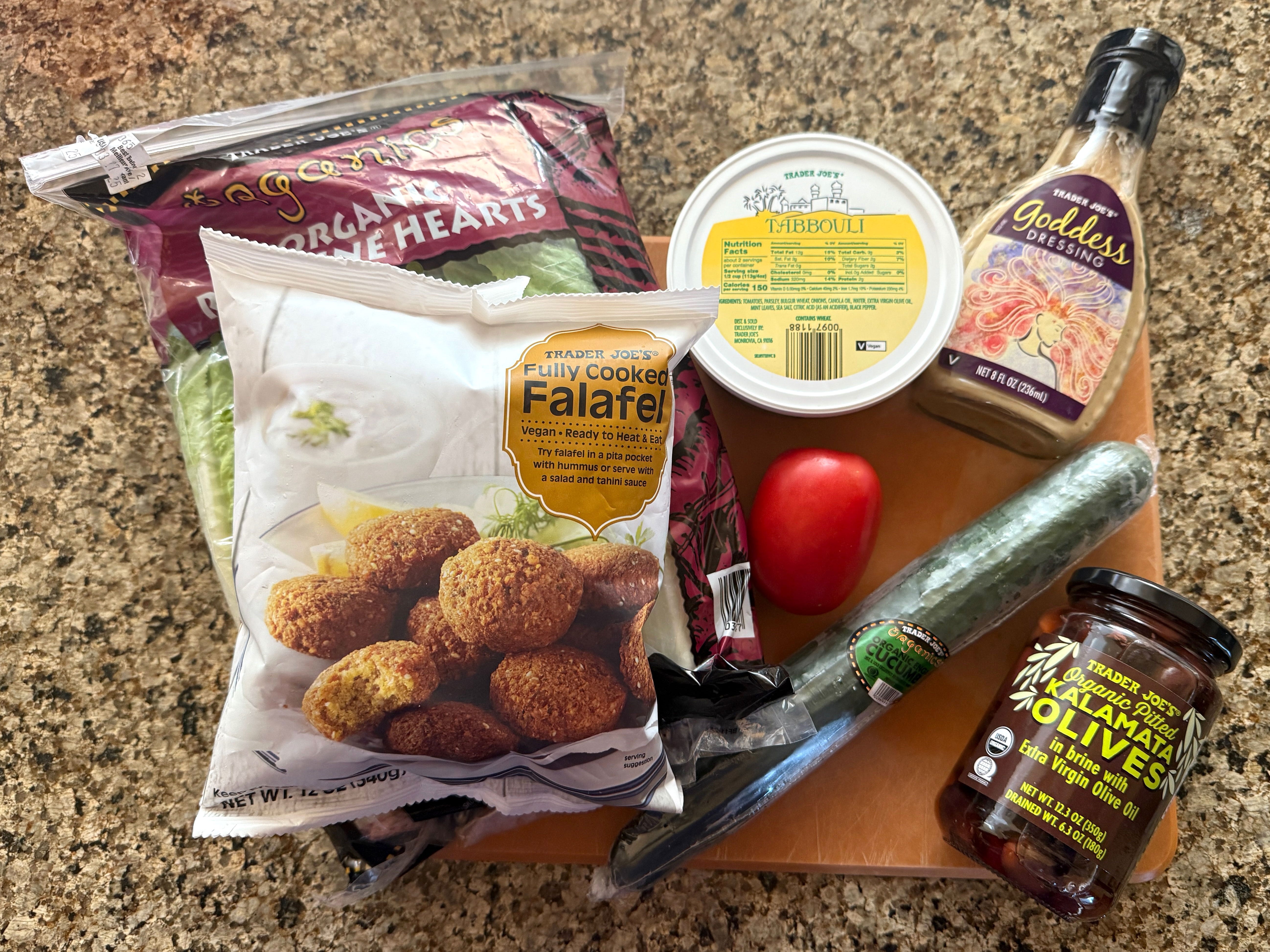 Romaine lettuce, a tomato, a cucumber, a jar of Trader Joe's pitted kalamata olives, a container of Trader Joe's Tabbouli, a bottle of Trader Joe's Goddess dressing, and a bag of frozen Trader Joe's falafel on a countertop.