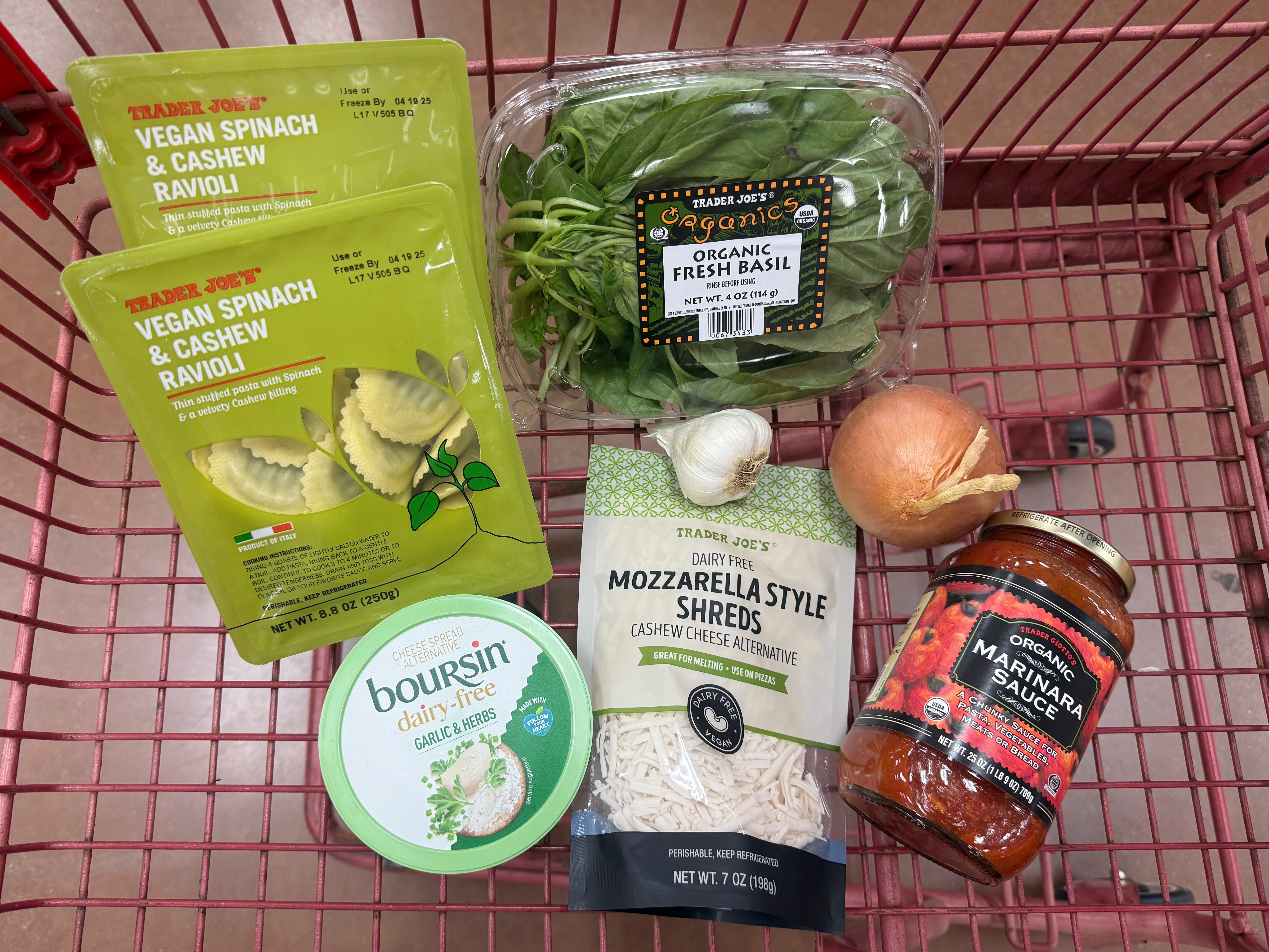 Two packages of Trader Joe's vegan ravioli, a package of fresh basil, garlic, an onion, a container of dairy-free Boursin cheese, a bag of vegan mozzarella shreds, and a jar of organic marinara sauce in a red shopping cart.
