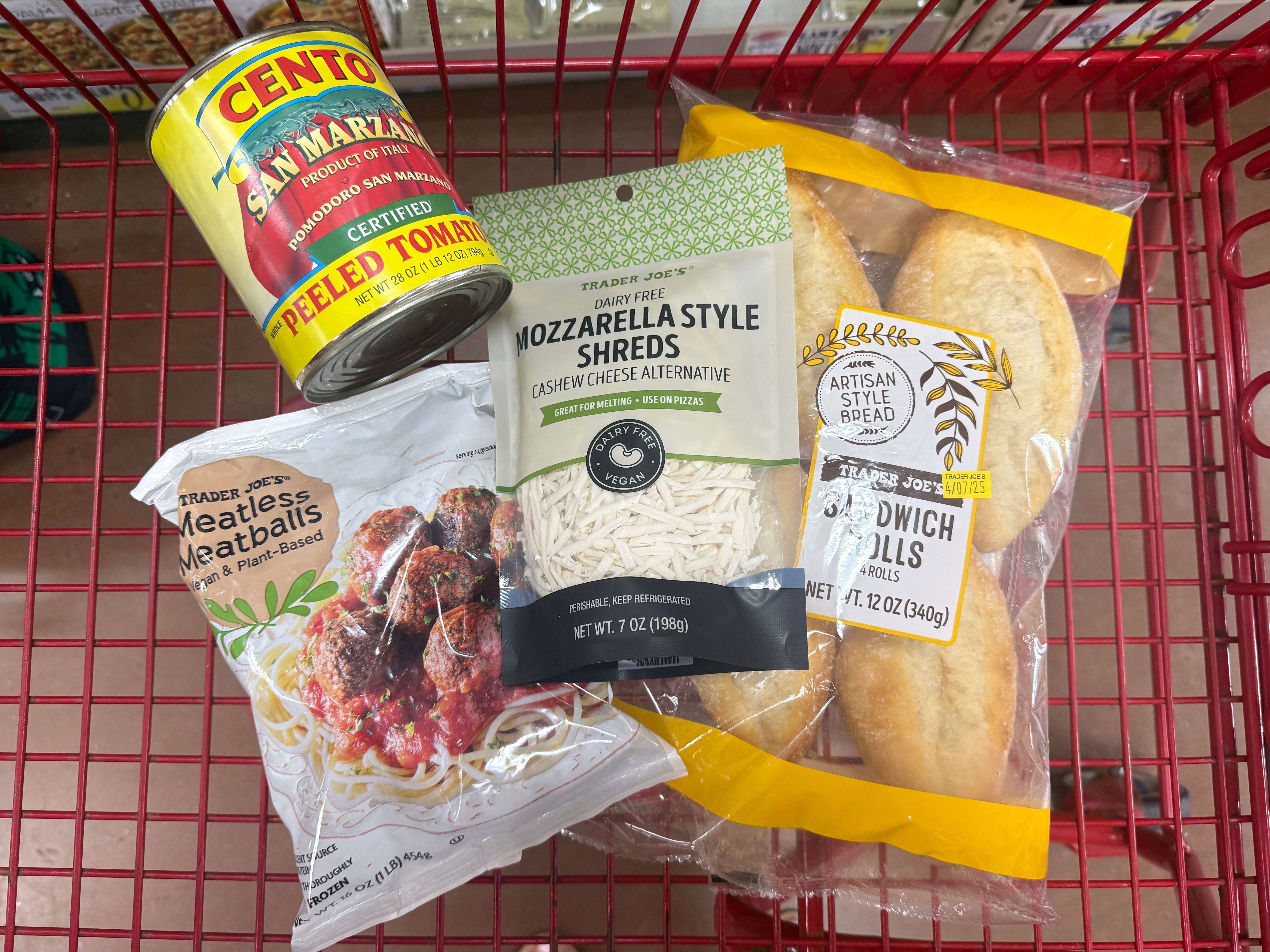 A can of San Marzano tomatoes, a bag of Trader Joe's vegan meatballs, a bag of dairy-free mozzarella shreds, and Trader Joe's sandwich rolls in a red shopping cart.