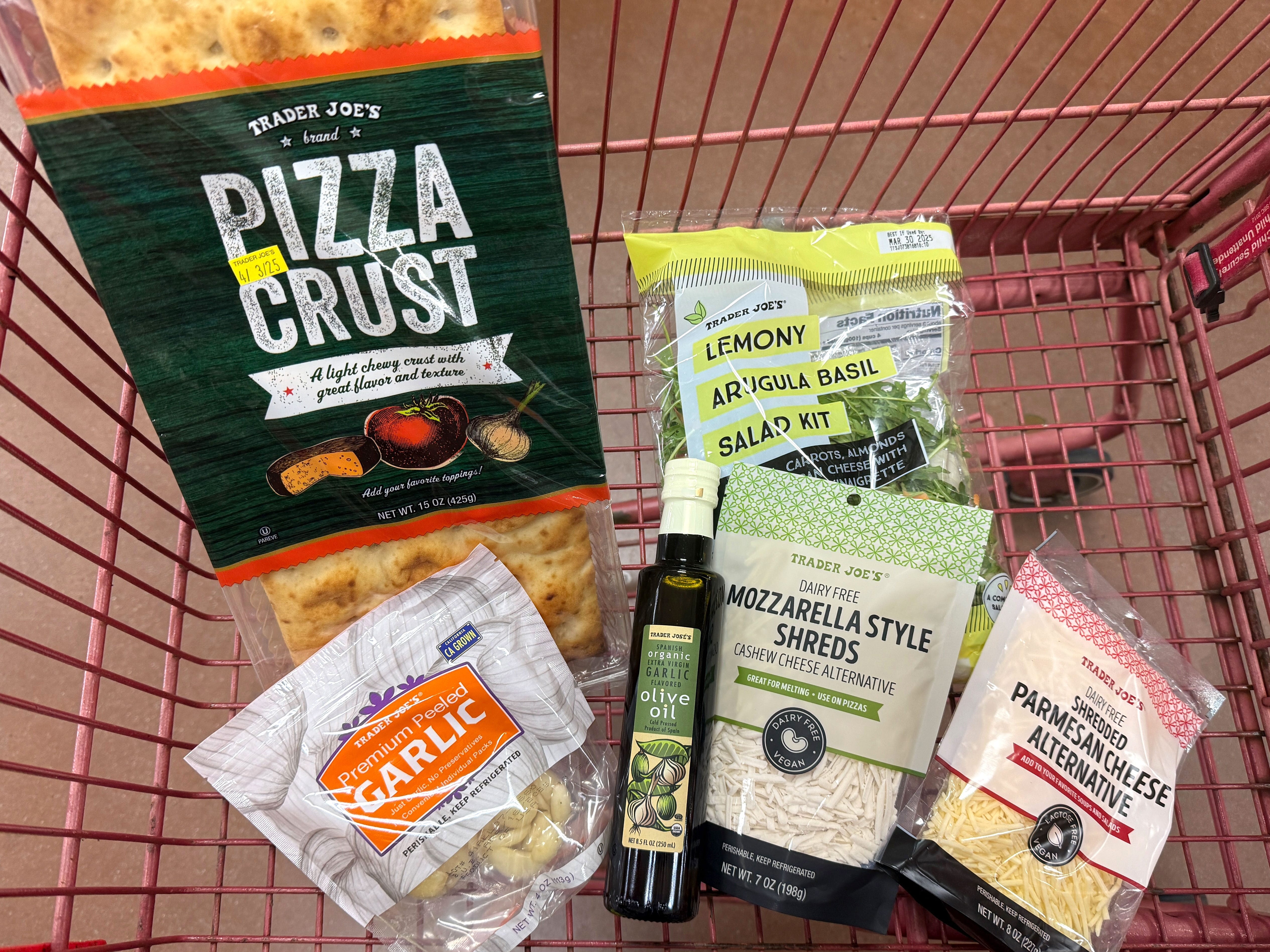 A bag of Trader Joe's pizza crust, a bag of peeled garlic, a bag of Trader Joe's lemony arugula basil salad, a bottle of olive oil, and bags of dairy-free mozzarella and Parmesan cheese in a red shopping cart.