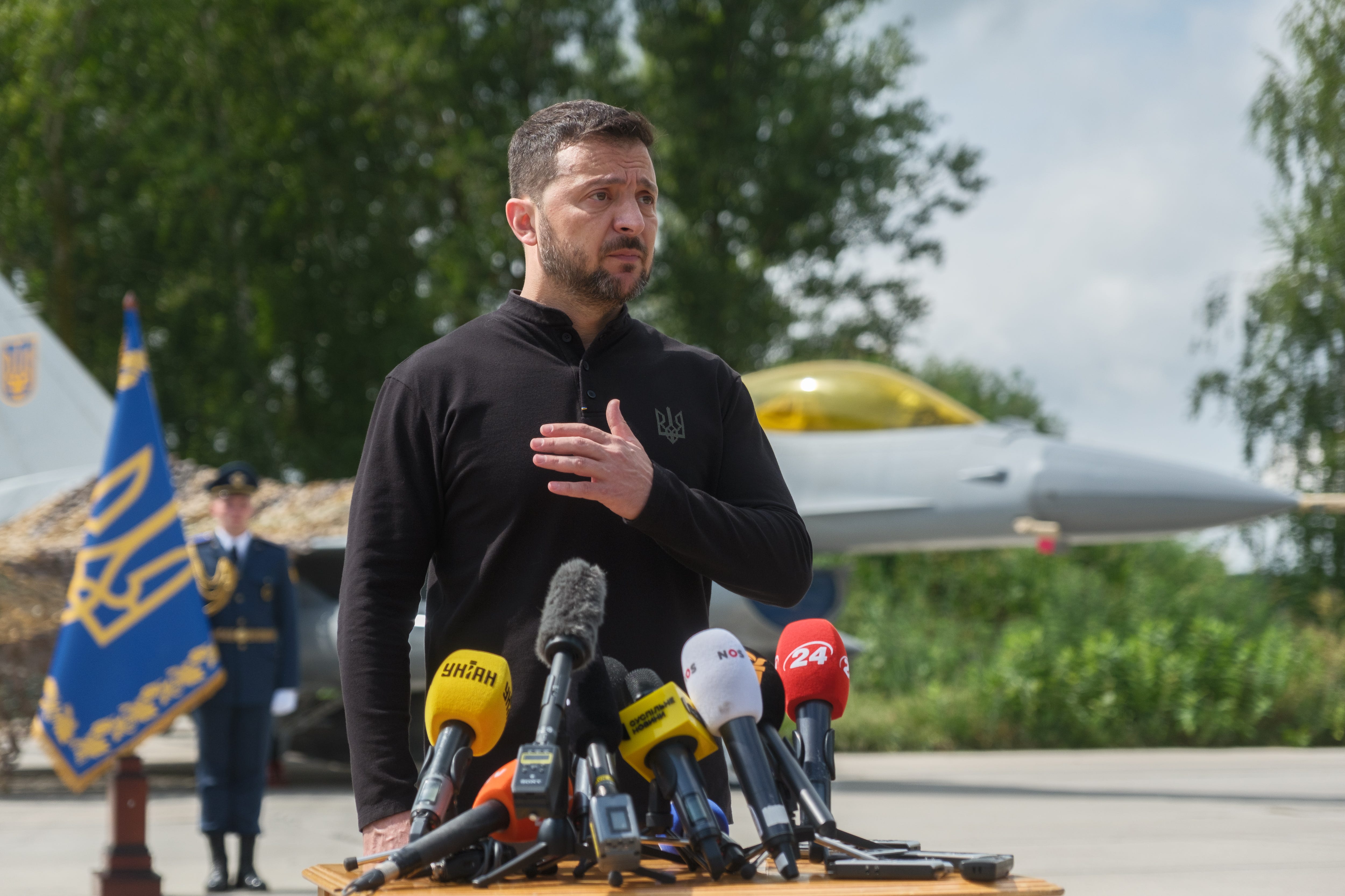 Ukrainian President Volodymyr Zelenskyi speaking into microphones with his hand over his chest. F-16 fighter jets are behind him.