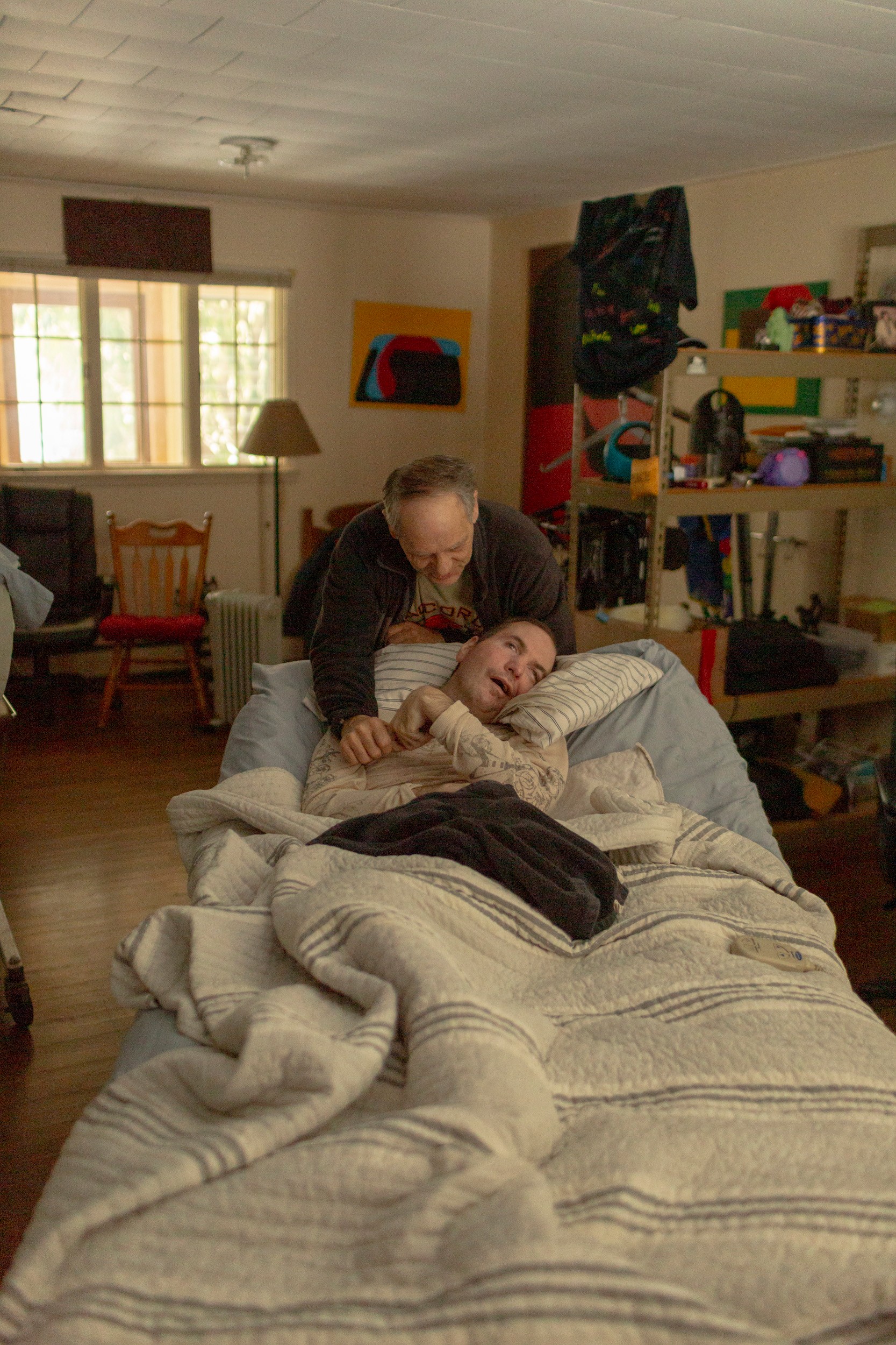 photo of man leaning over head of hospital bed and holding hand of the man lying in it in home living room