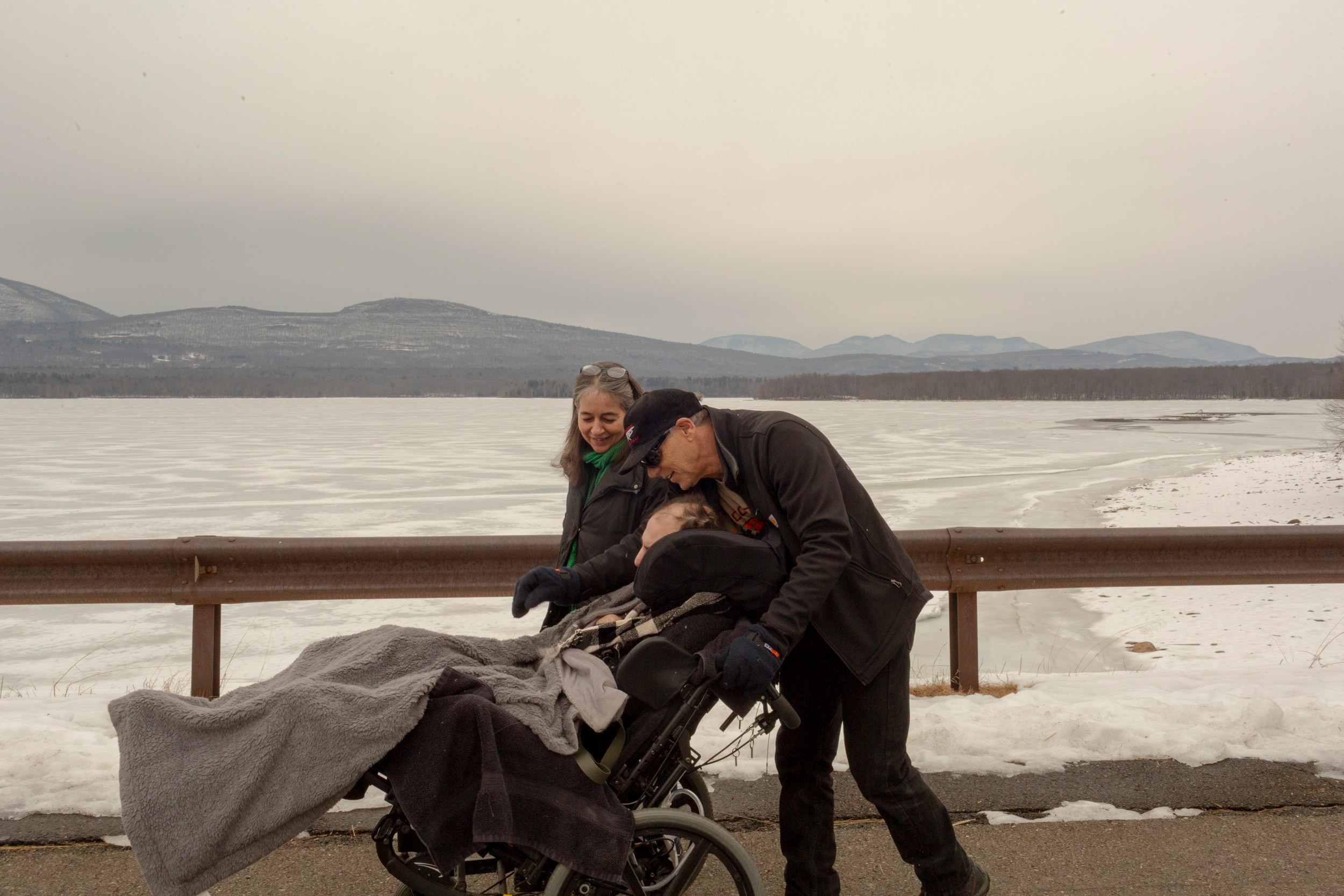 photo of woman and man smiling and walking outdoors pushing brother in wheelchair, the man bending over head of his brother, with snowy landscape and mountains behind
