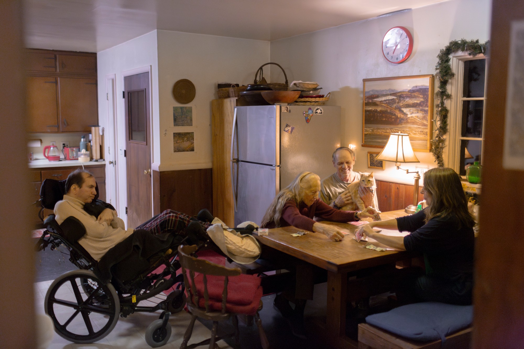 photo of 3 people playing cards at warmly lit kitchen table with man in wheelchair next to head of table