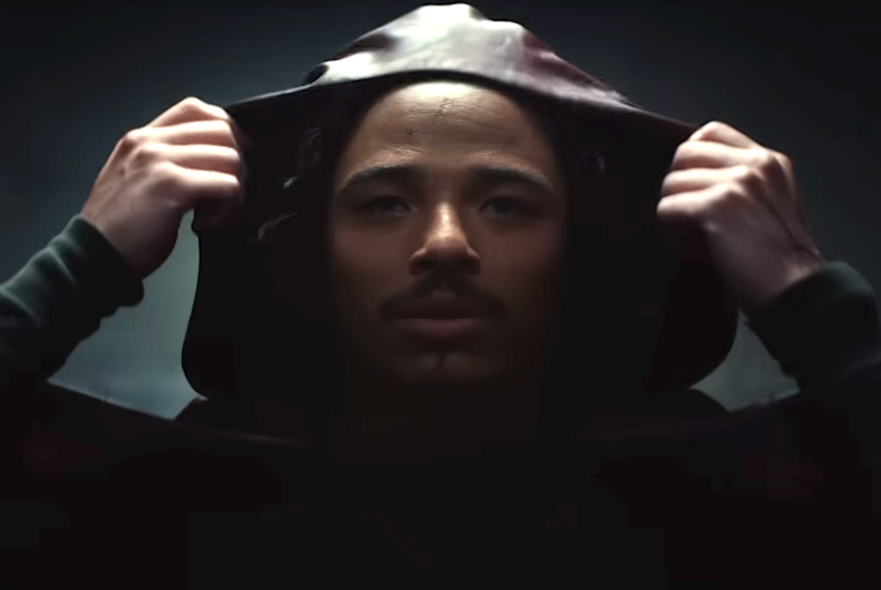 A man with dark hair and a goatee is pulling a dark red hood over his head.