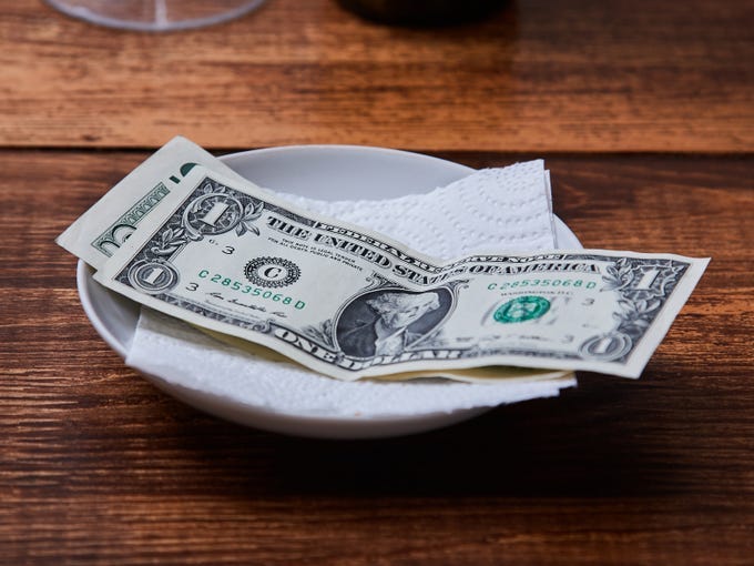 A white plate with a napkin and a $5 bill and $1 bill stacked on top of it