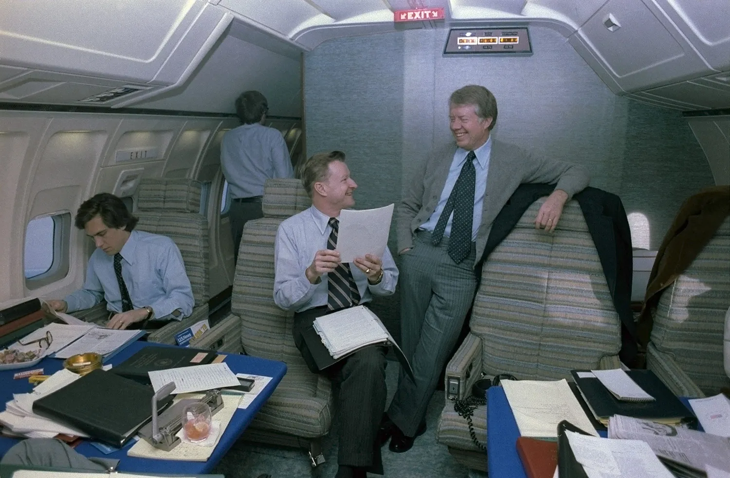 Jimmy Carter stands leaning on a plane seat while he smiles and talks with a seated Zbigniew Brzezinski who holds papers. At left is a staffer with other papers.