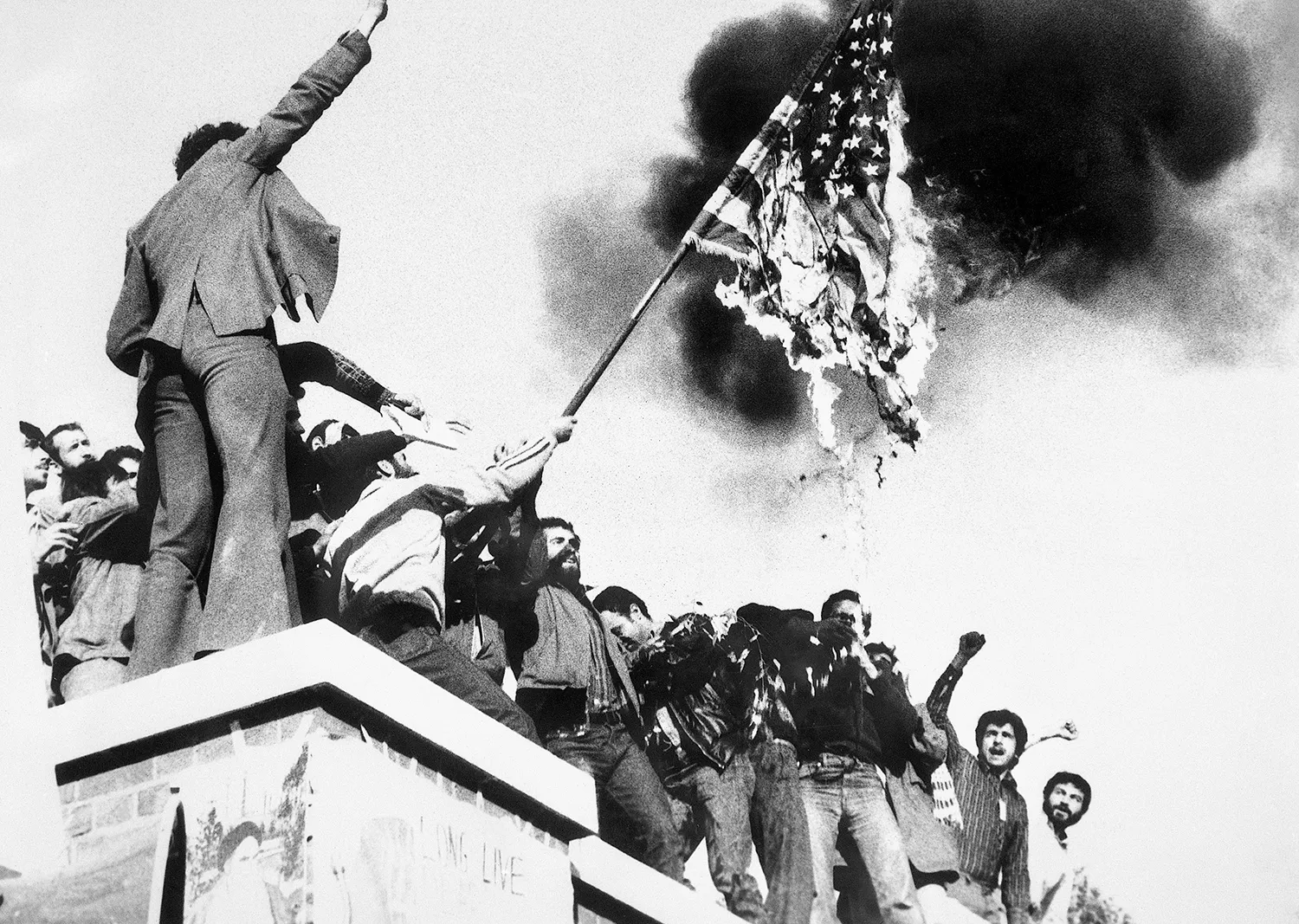 A black and white photo shows demonstrators with fists in the air as they stand atop a building burning an American flag.