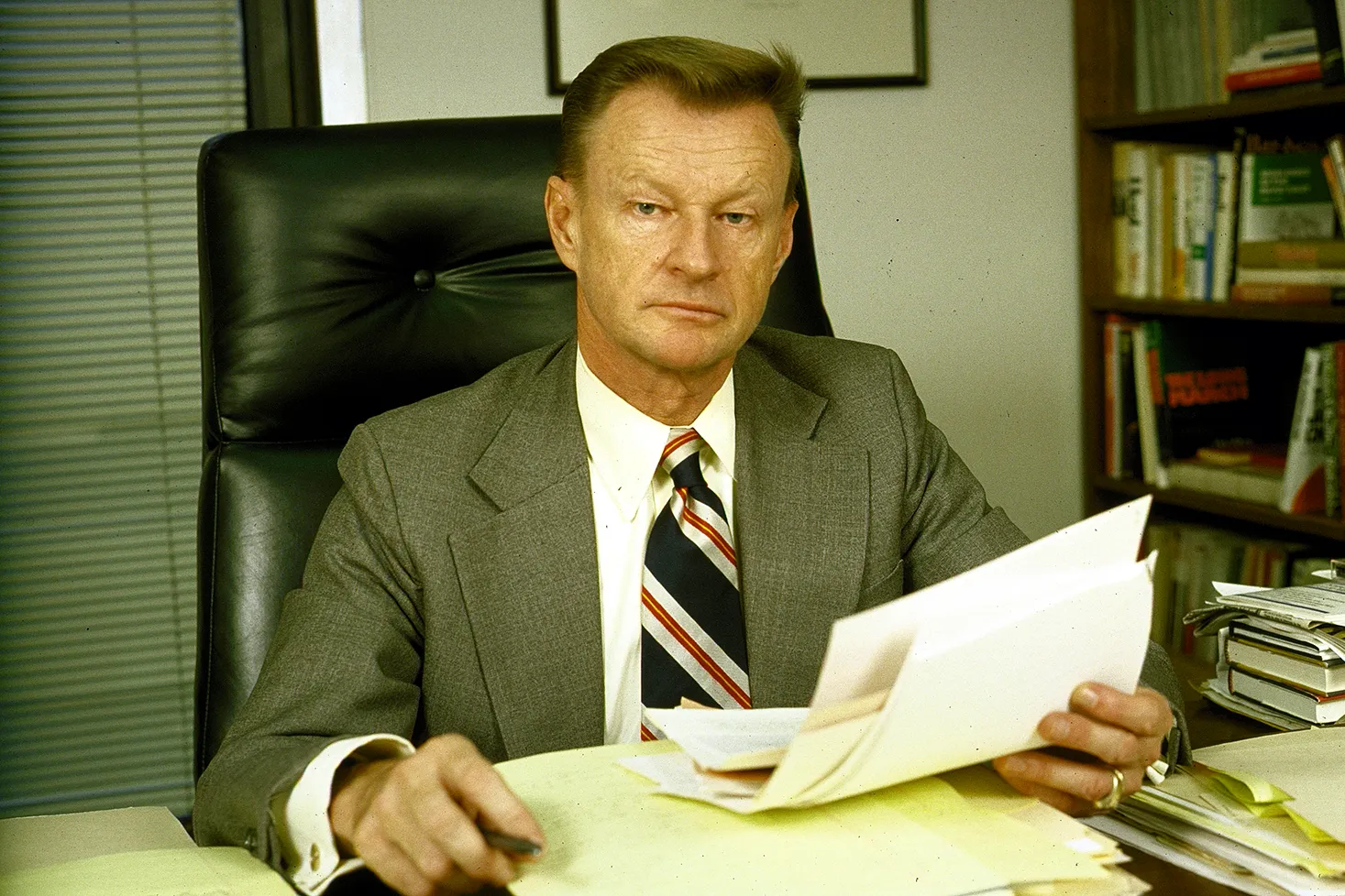 A man in a tie holds papers while he sits a desk in an office.