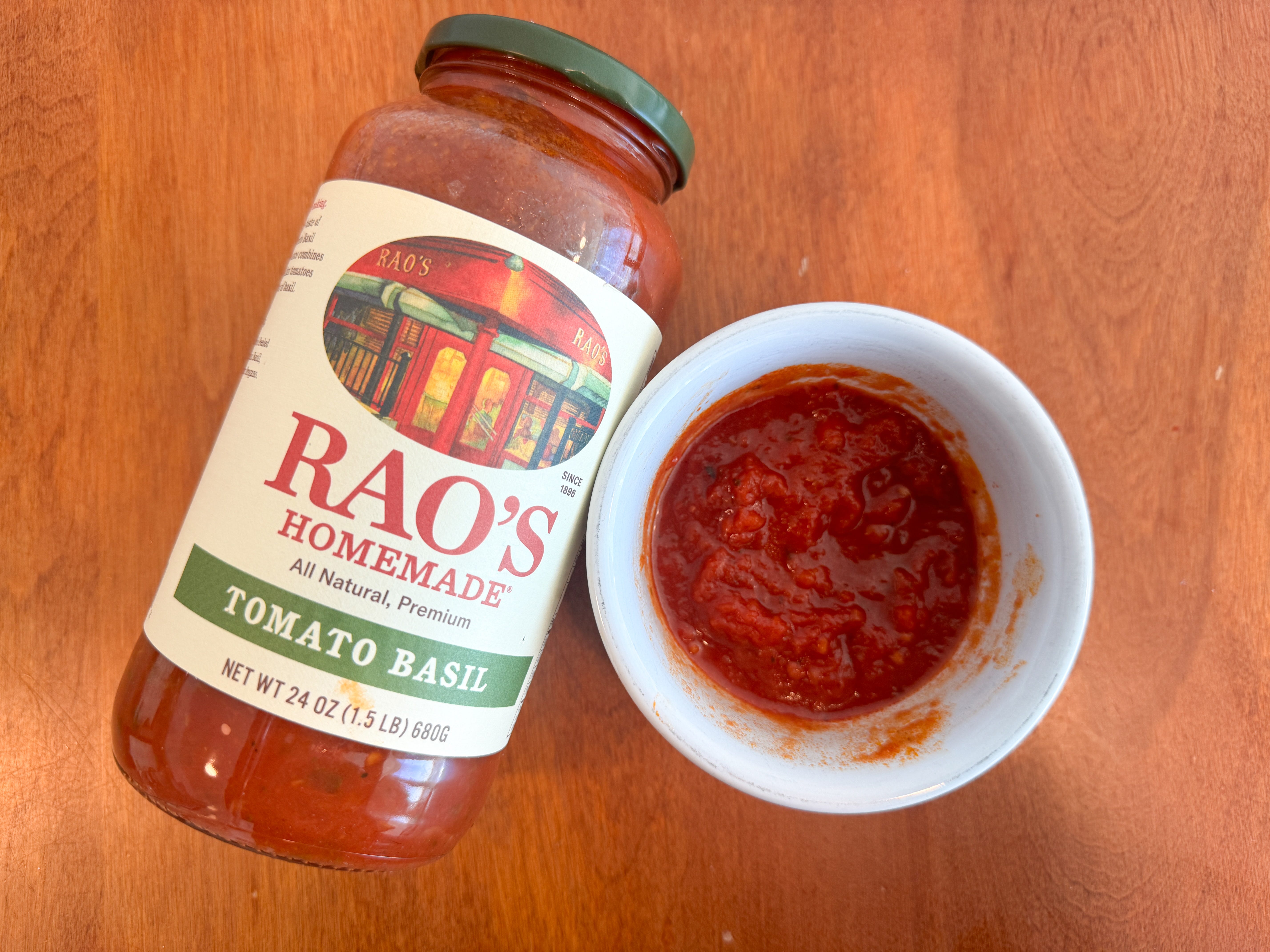 A small bowl of red tomato sauce next to a jar of Rao's Homemade tomato-basil pasta sauce lying on a wooden table.