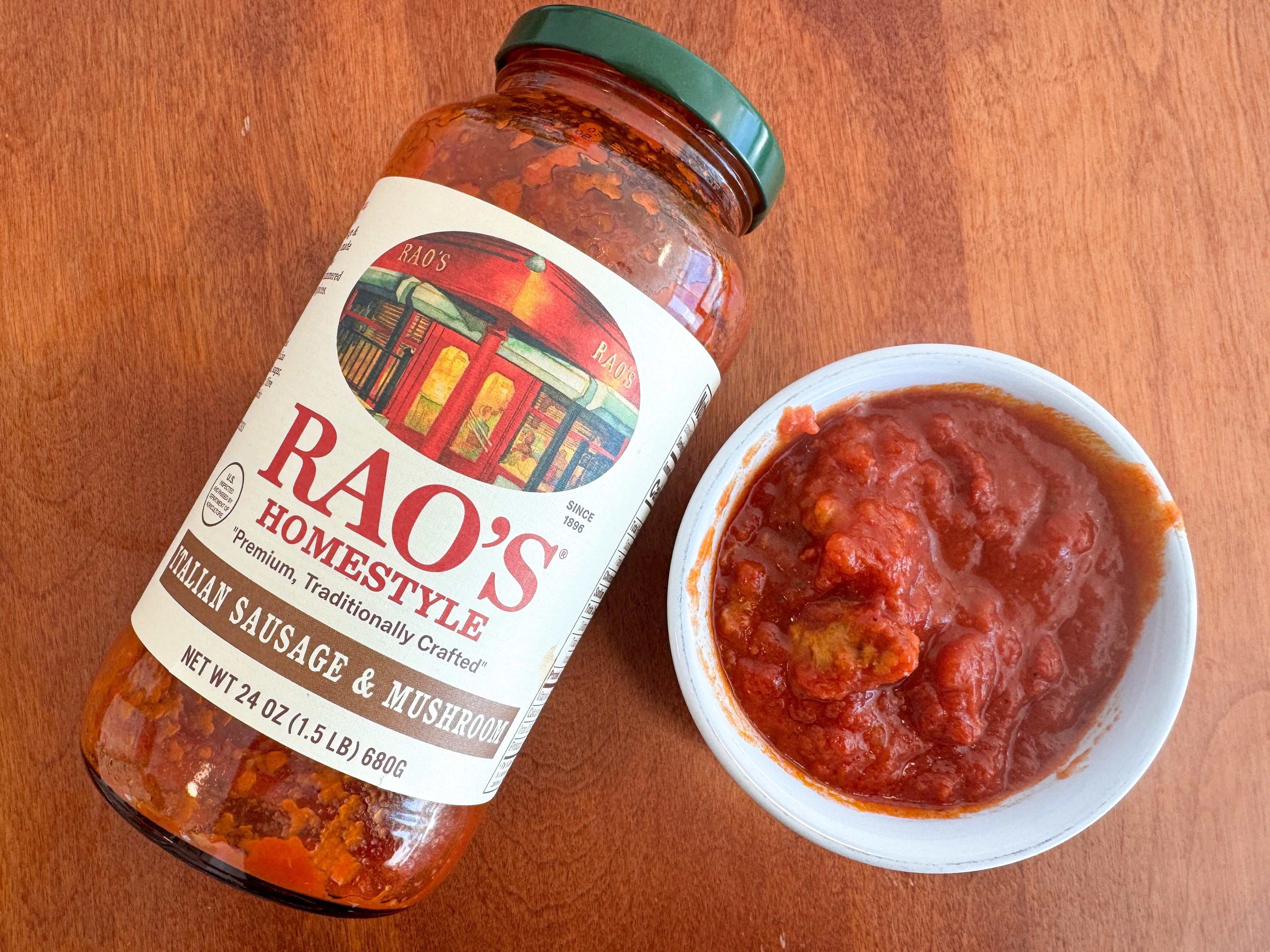 A small bowl of red tomato sauce next to a jar of Rao's Homemade Italian-sausage-and-mushroom pasta sauce lying on a wooden table.