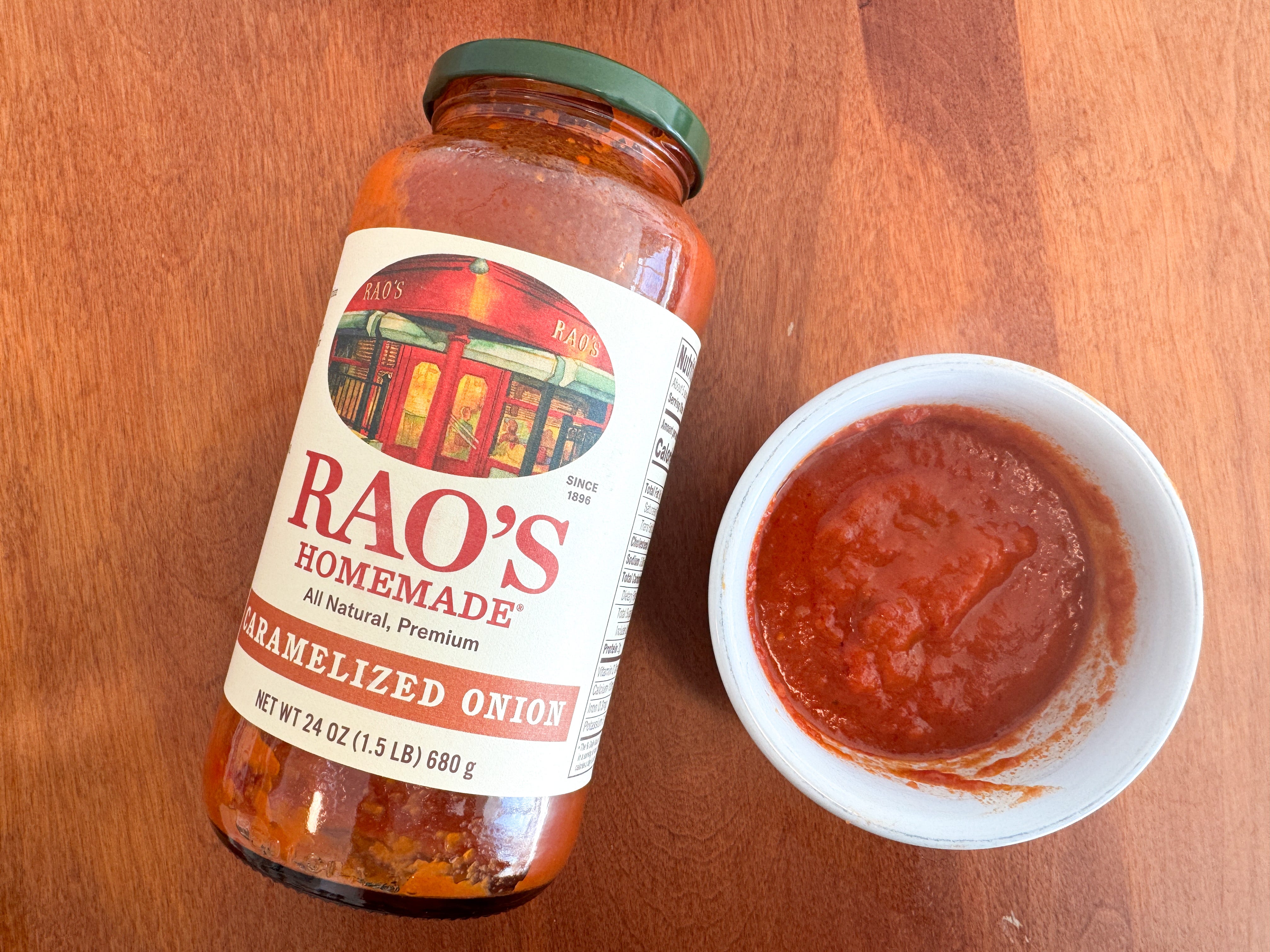 A small bowl of red tomato sauce next to a jar of Rao's Homemade carmelized-onion pasta sauce lying on a wooden table.