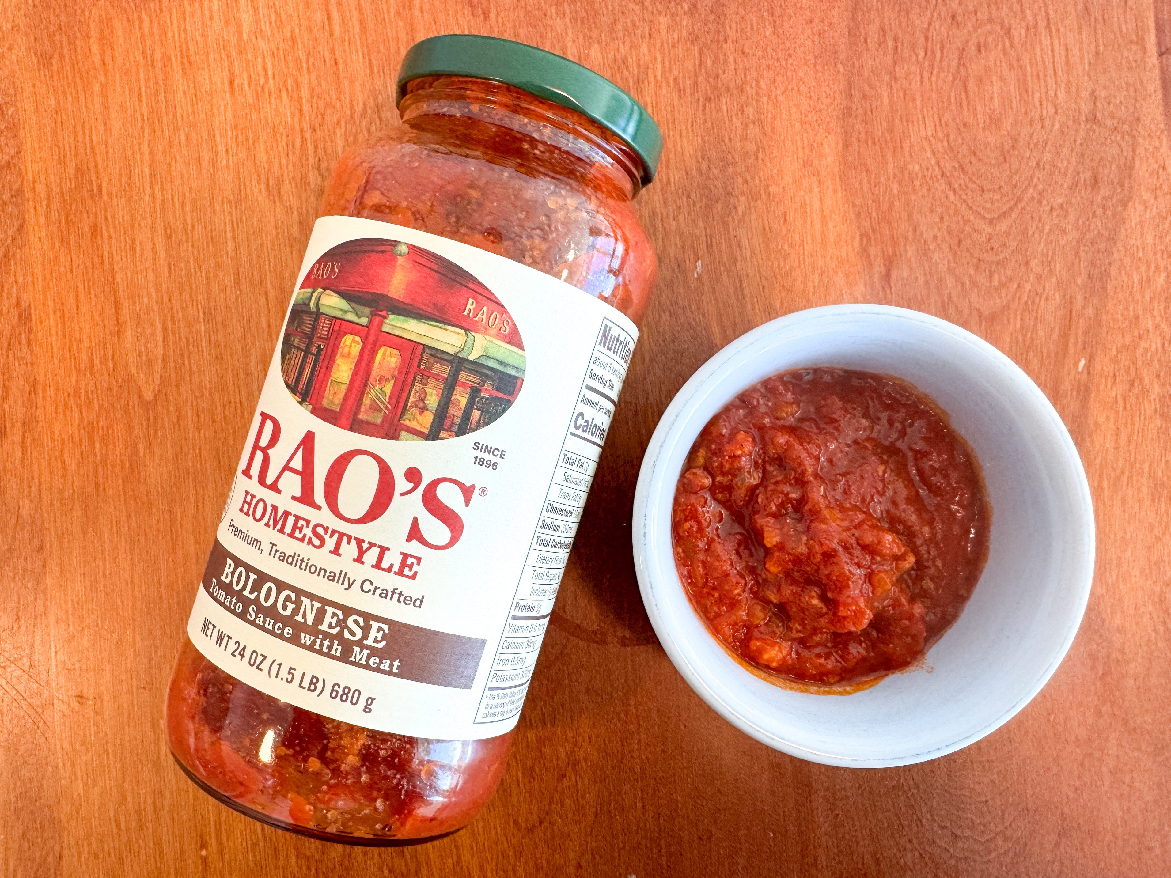 A small bowl of red tomato sauce next to a jar of Rao's Homemade bolognese pasta sauce lying on a wooden table.