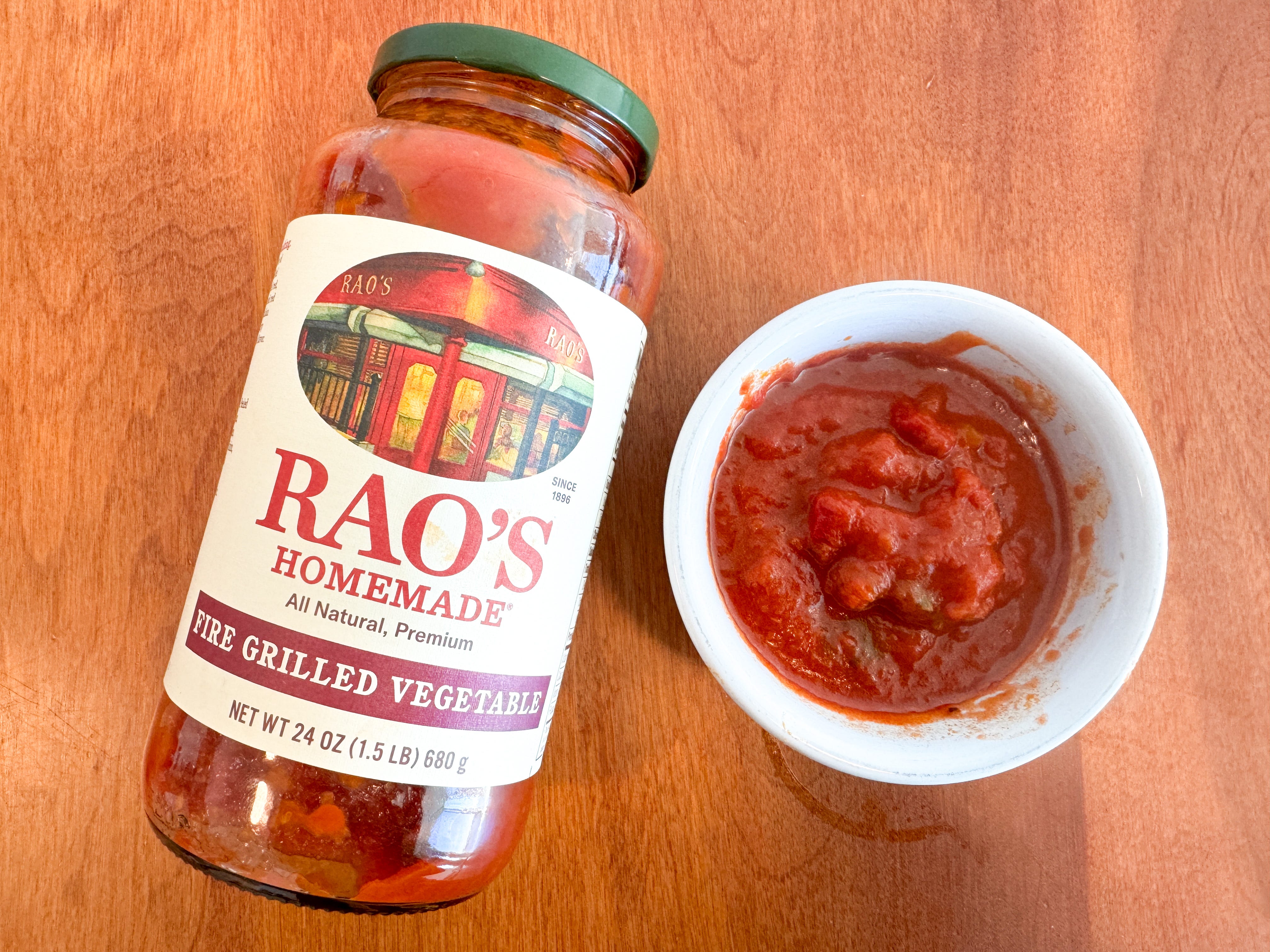 A small bowl of red tomato sauce next to a jar of Rao's Homemade fire-grilled-vegetable pasta sauce lying on a wooden table.
