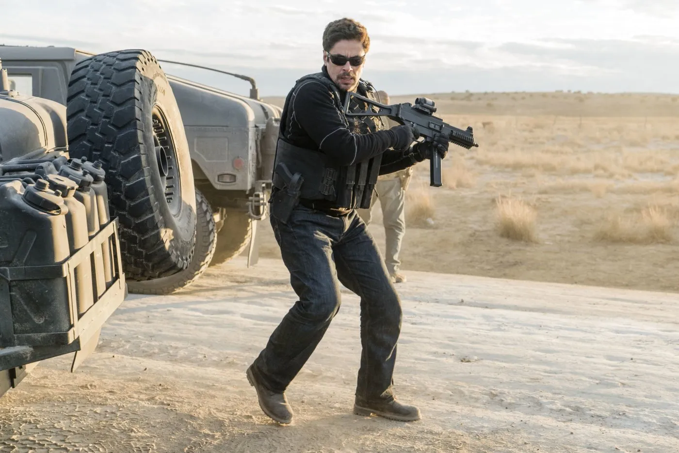 A man with a gun patrols with a weapon drawn at the back of a car on a dirt road.