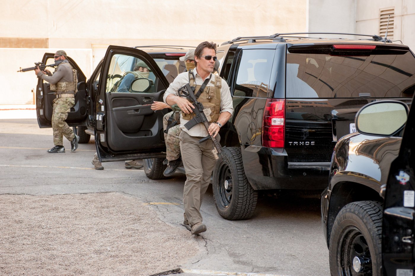 A man in police gear with a large gun walks walks purposefully out of a line of cars on a dirt road.
