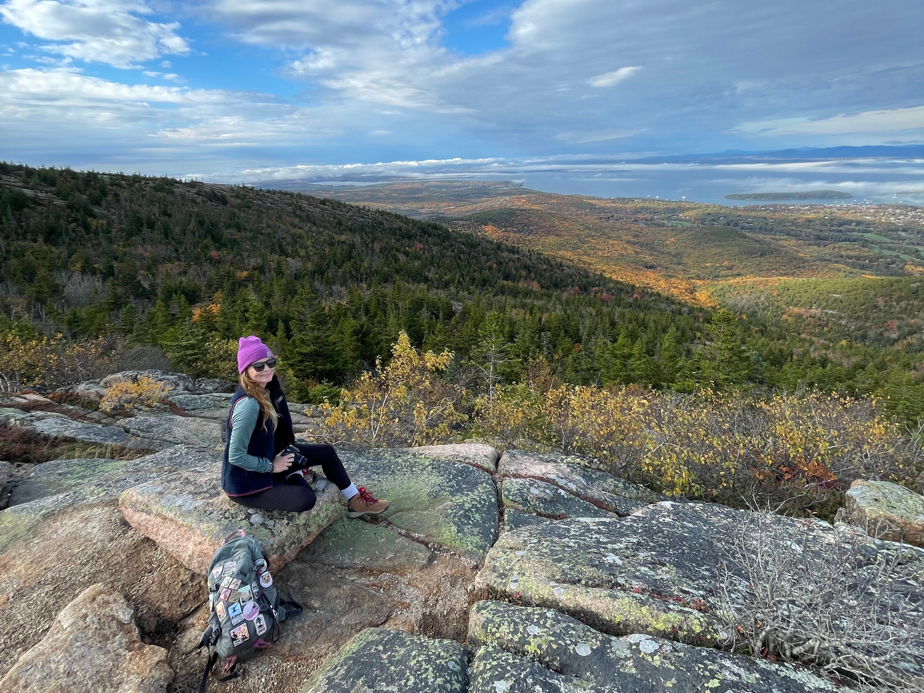 A woman sitting on a rock formation with her backpack next to her on a cloudy day. Behind her are mountains covered with trees.