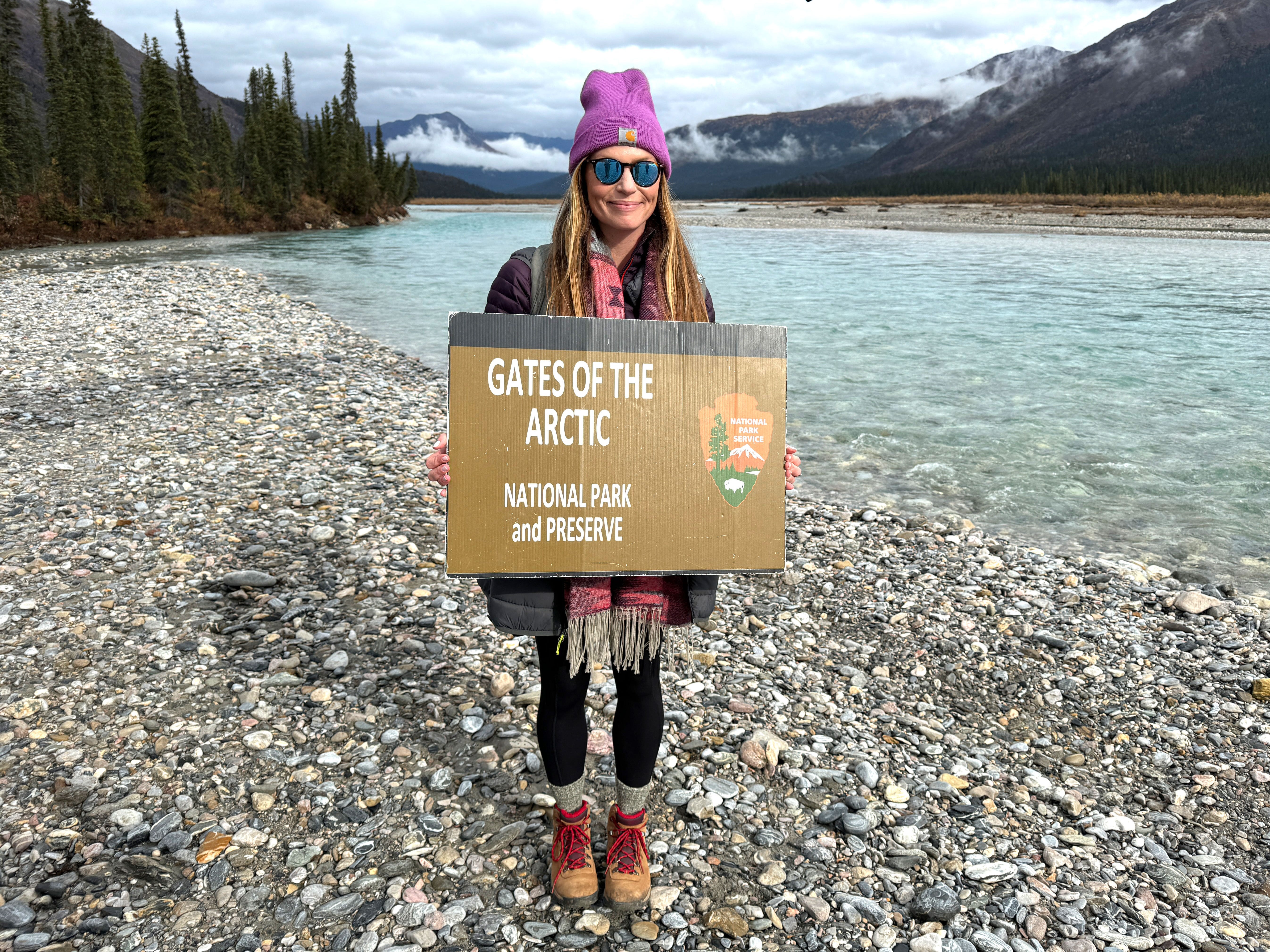 The author stands on a rocky beach with trees, mountains, and a lake in the background. She is holding a cardboard sign that reads, "Gates of the Arctic National Park and Preserve."