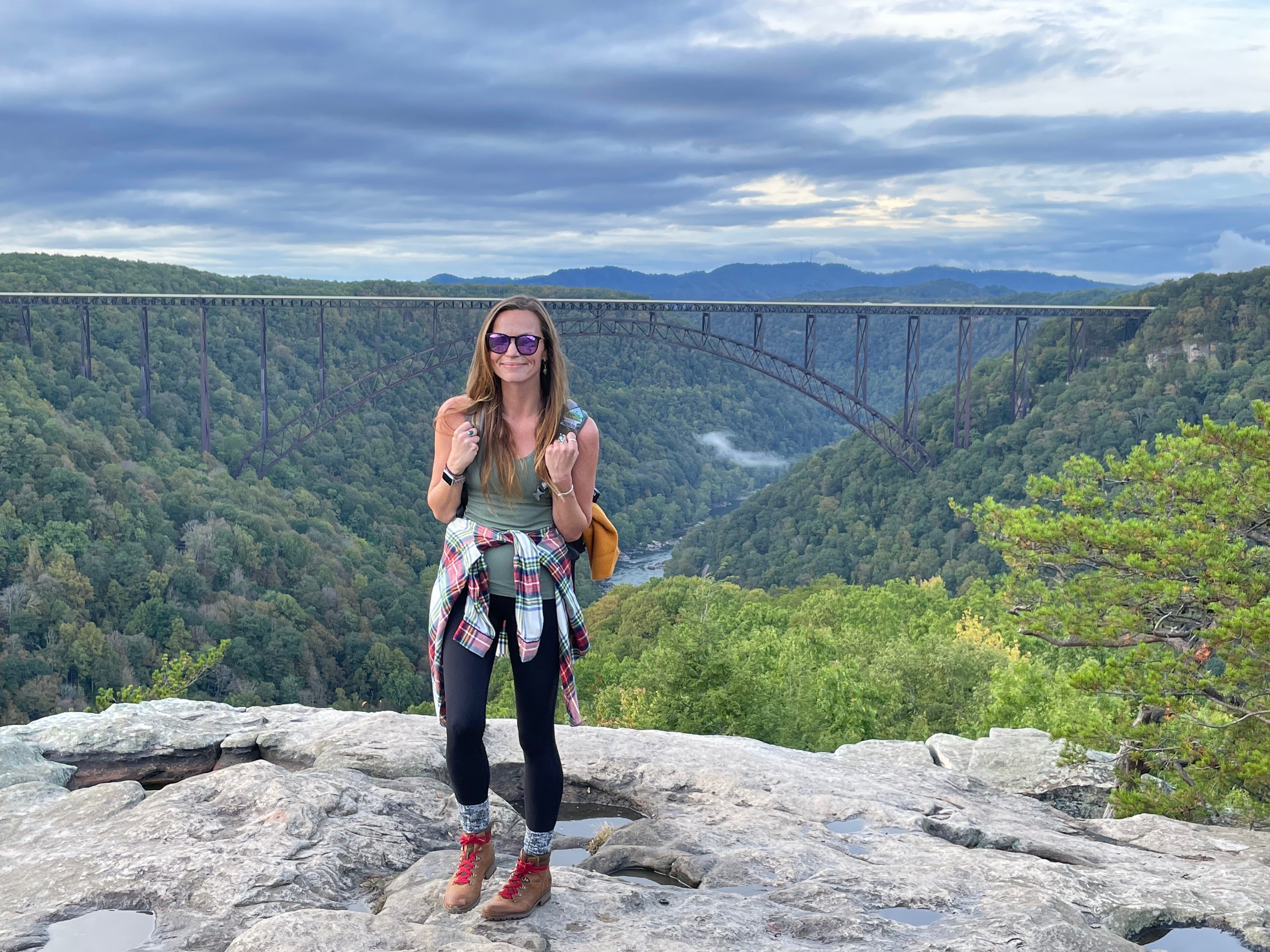 A woman standing on a rock in front of a large bridge with tree-covered mountains.