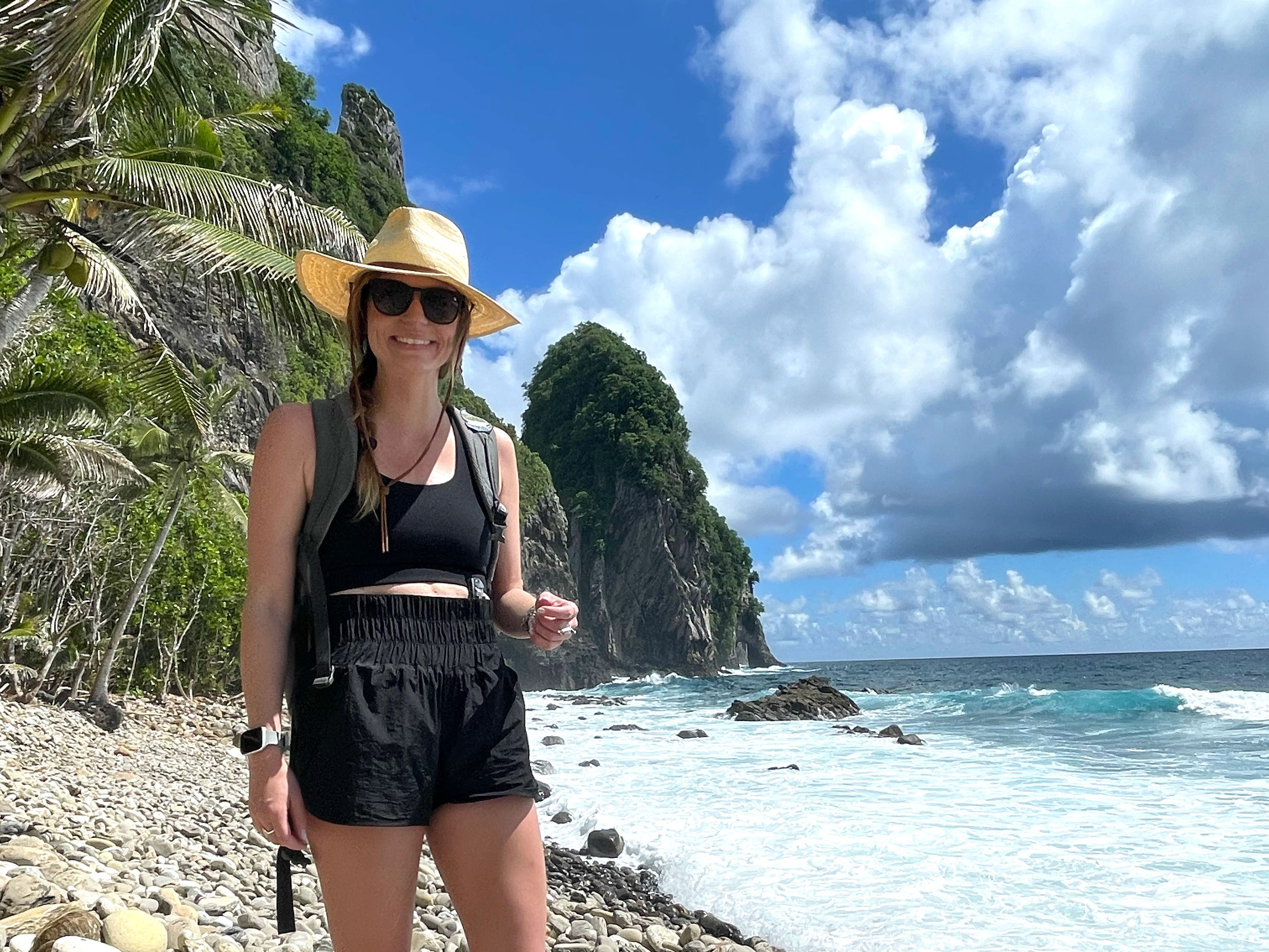 The author stands on a rocky beach with large trees and rock formations behind her.