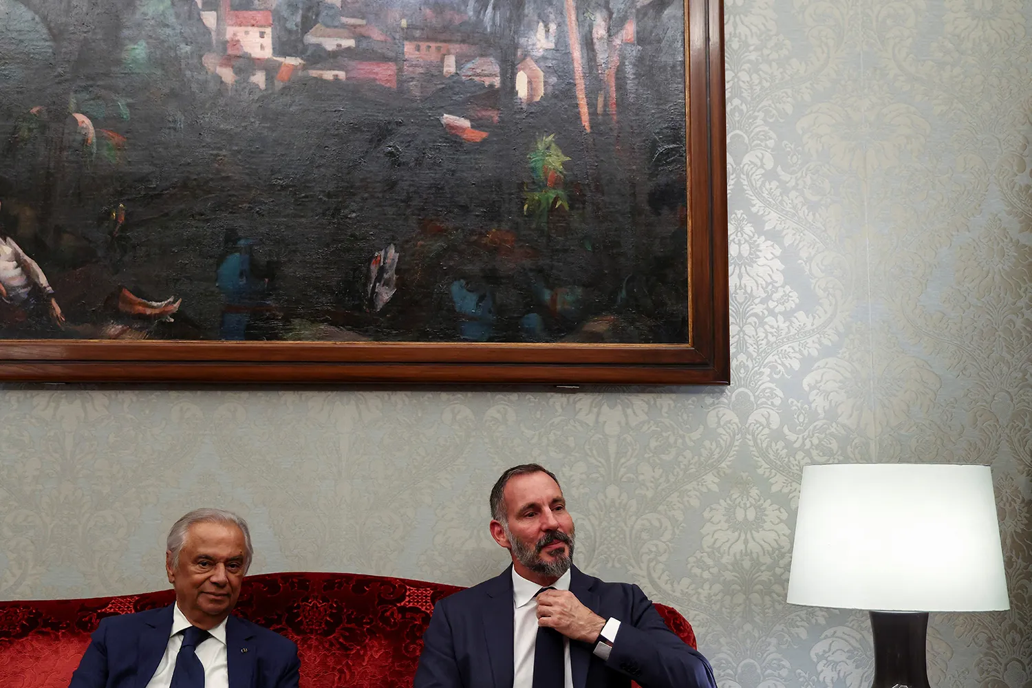 Two men in suits sit on a red couch next to a lamp. The corner of a massive oil landscape painting is seen hanging on the wall over their heads. The man on the right, the Aga Khan, has his hand raised to adjust his tie.