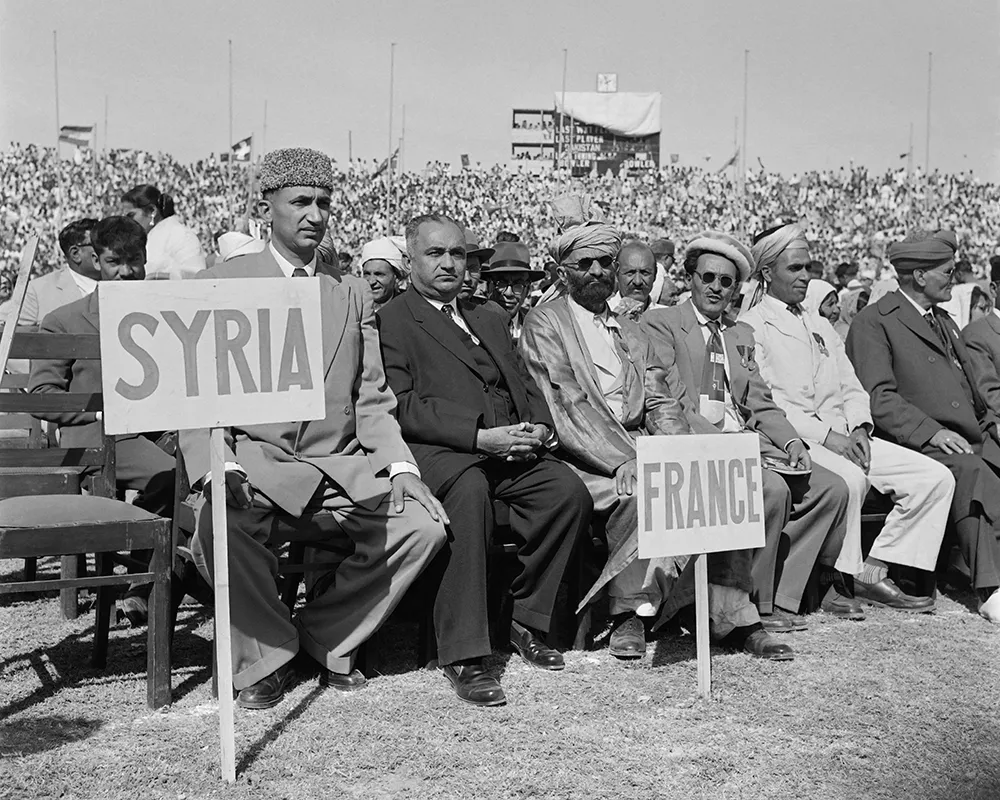 In this black-and-white photo, signs for Syria and France stand on picket poles are stuck into the round in front of a large crowd of people seated in an outdoor space.