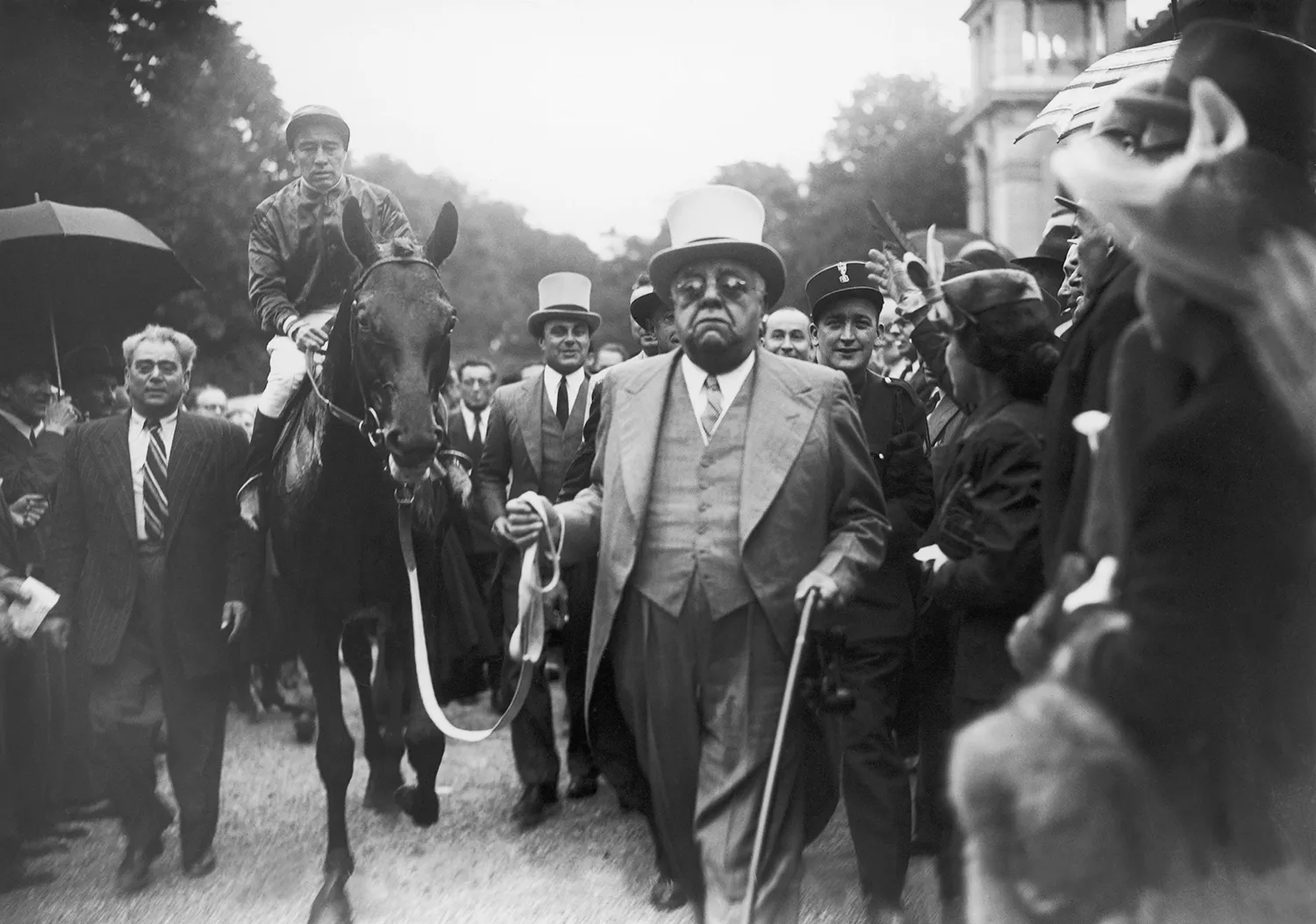 In this black-and-white photo, a heavyset man in a three-piece suit, glasses, and top hat walks with a cane as he holds the lead of a racehorse ridden by a jockey as a crowd gathers around on either side of the path they walk down.