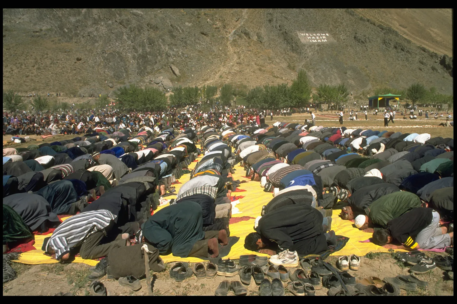 Rows of men are seen kneeling on prayer mats with their foreheads on the ground in prayer. Behind them is a large crowd and a mountainside.