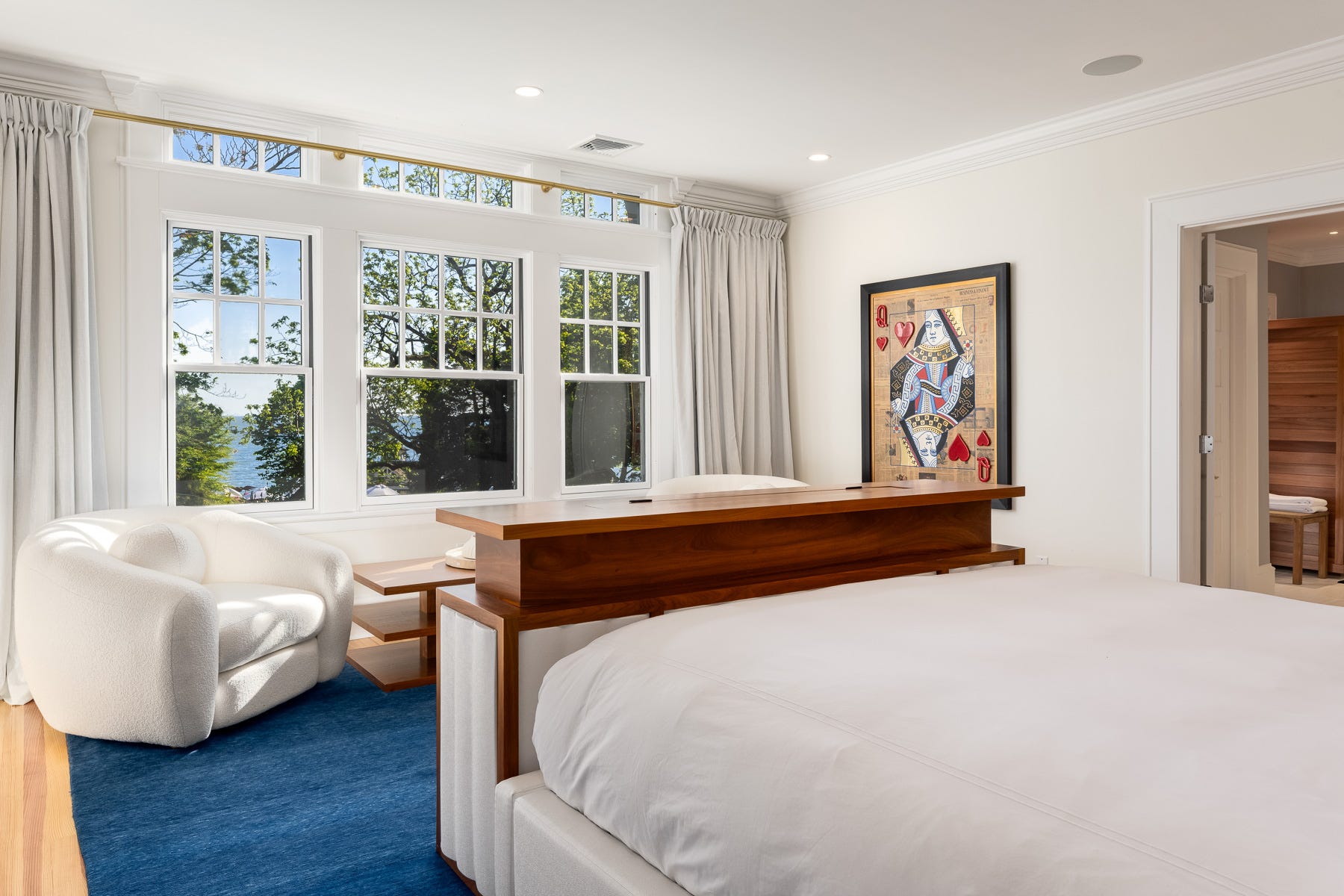 A bedroom with blue carpet and a painting of a playing card on the wall, and windows overlooking the water.