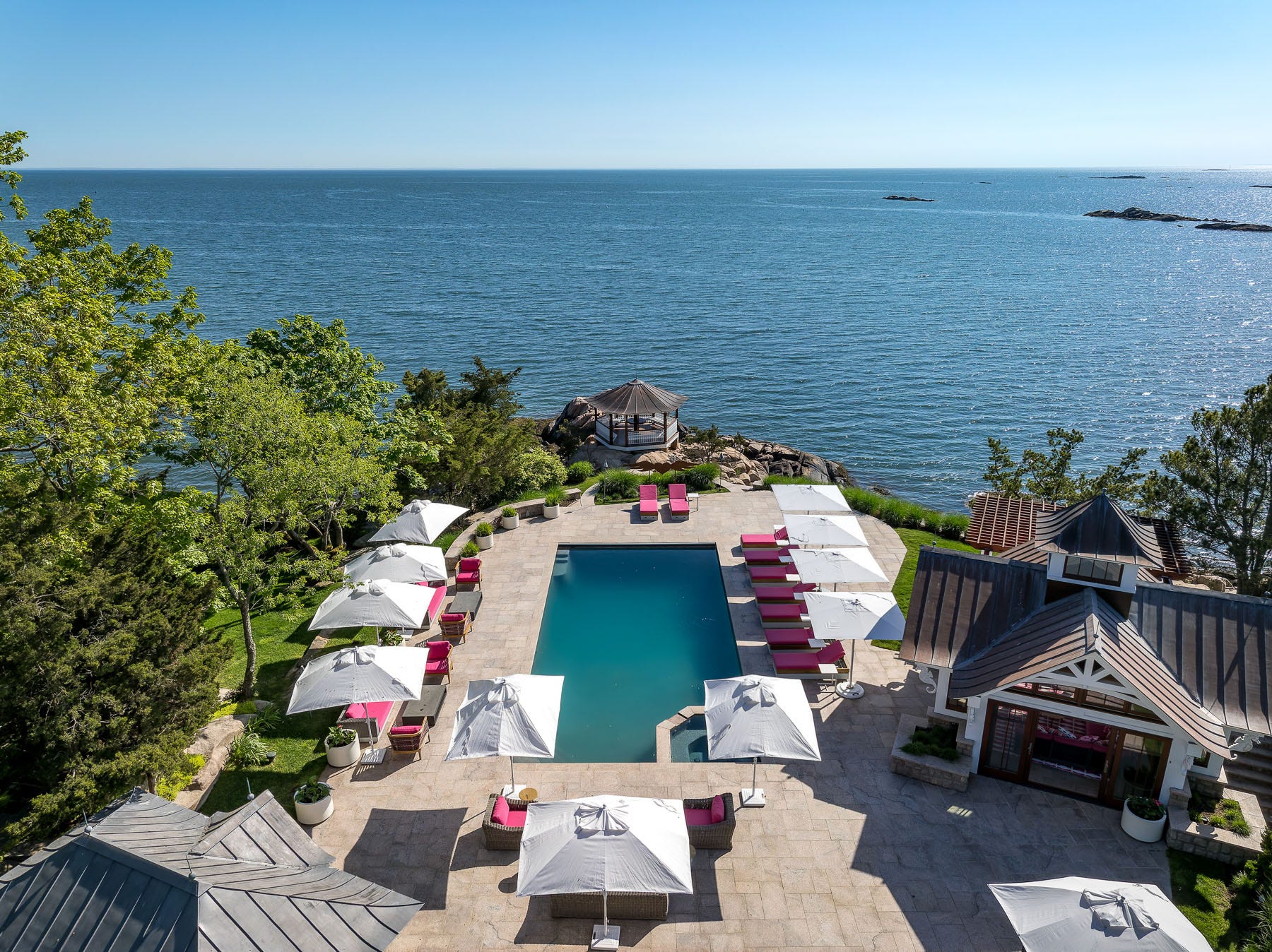 A waterfront pool with pink deck chairs and white umbrellas, as well as a gazebo looking out on Long Island Sound.